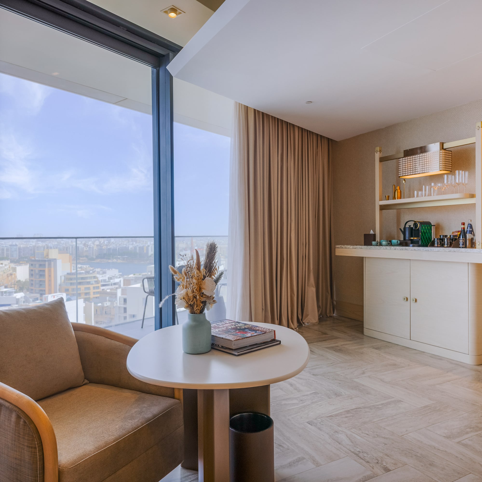 Modern hotel suite lounge with city view, armchair, and coffee bar.