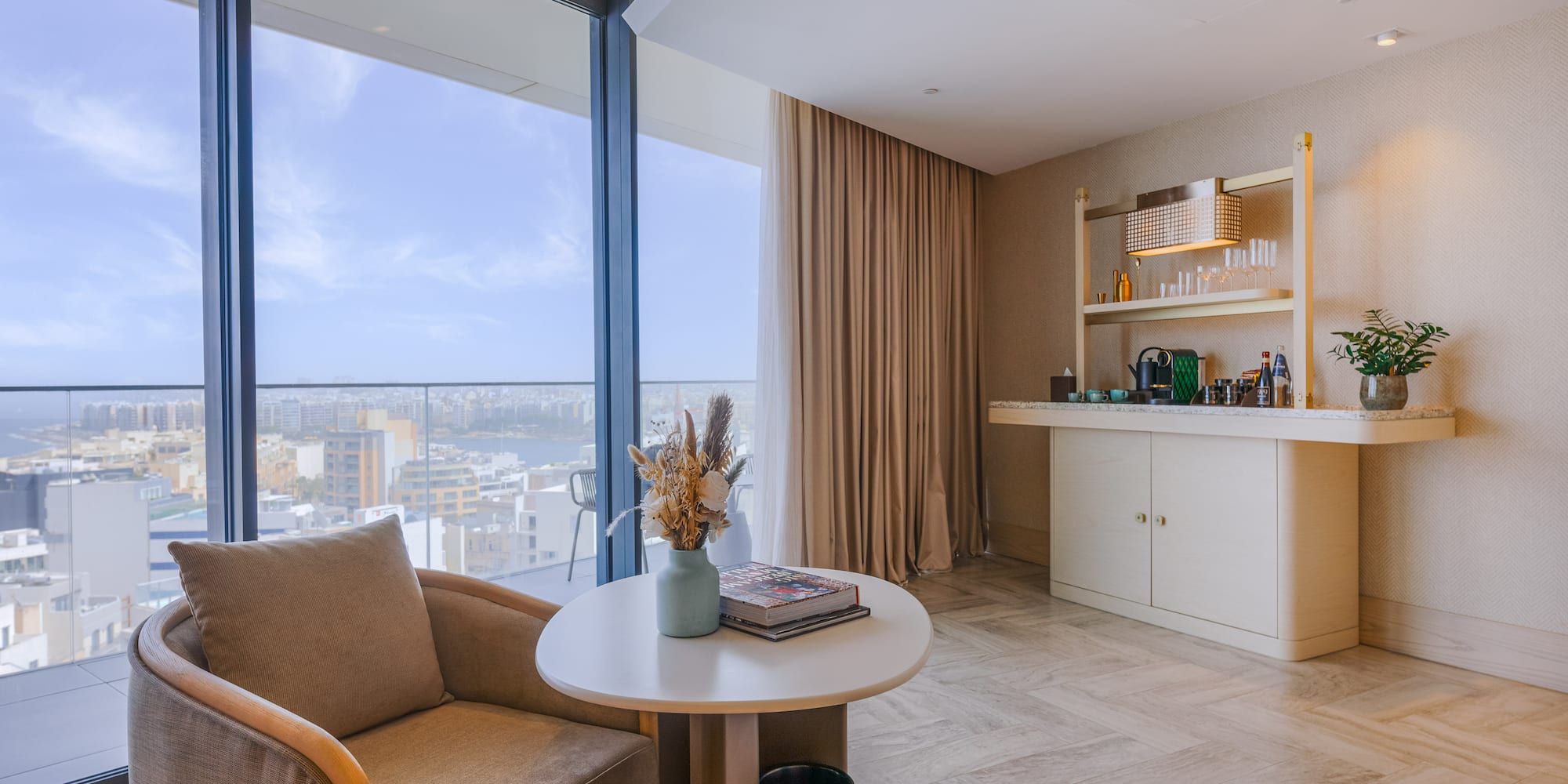 Modern hotel suite lounge with city view, armchair, and coffee bar.