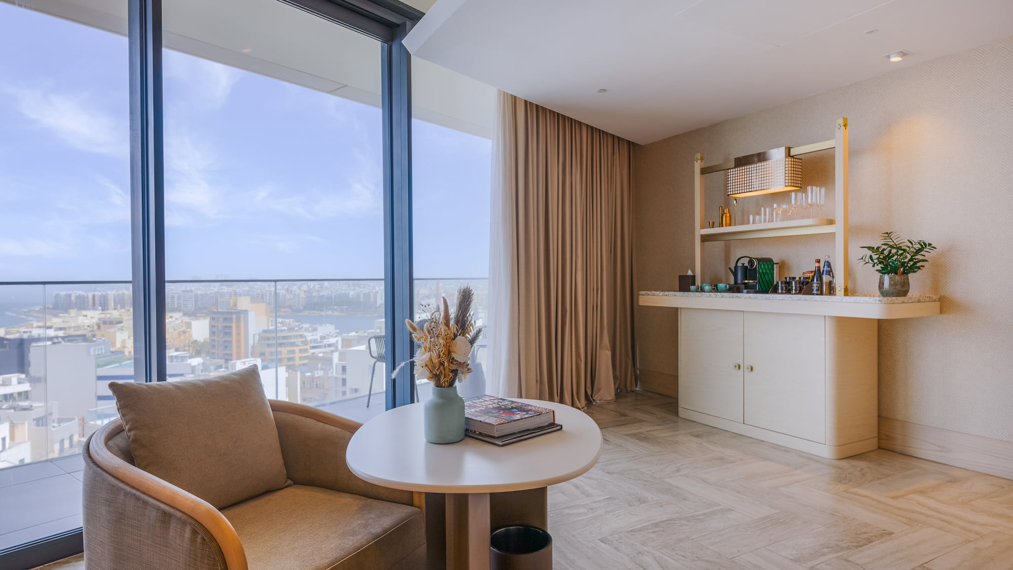Modern hotel suite lounge with city view, armchair, and coffee bar.