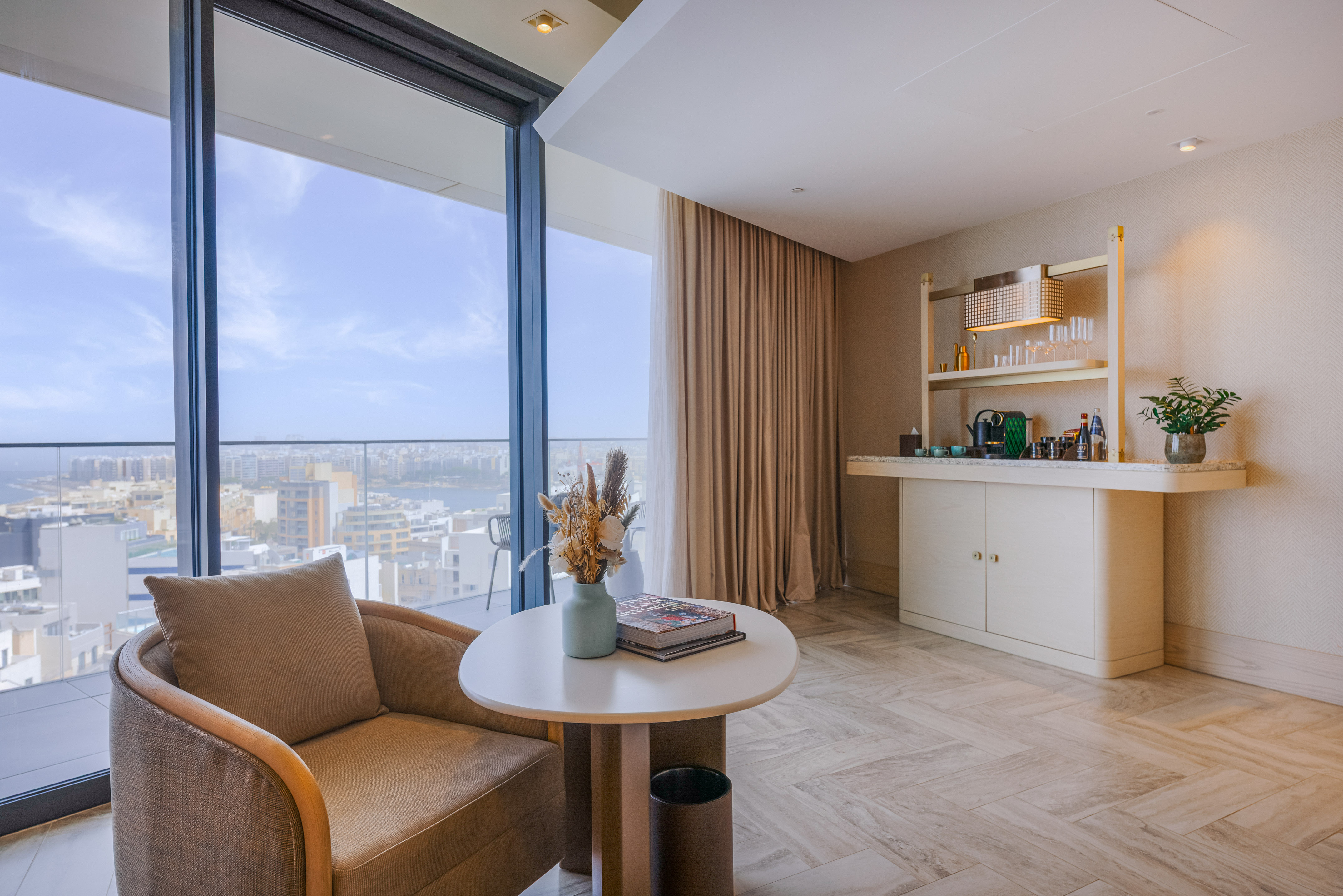 Modern hotel suite lounge with city view, armchair, and coffee bar.