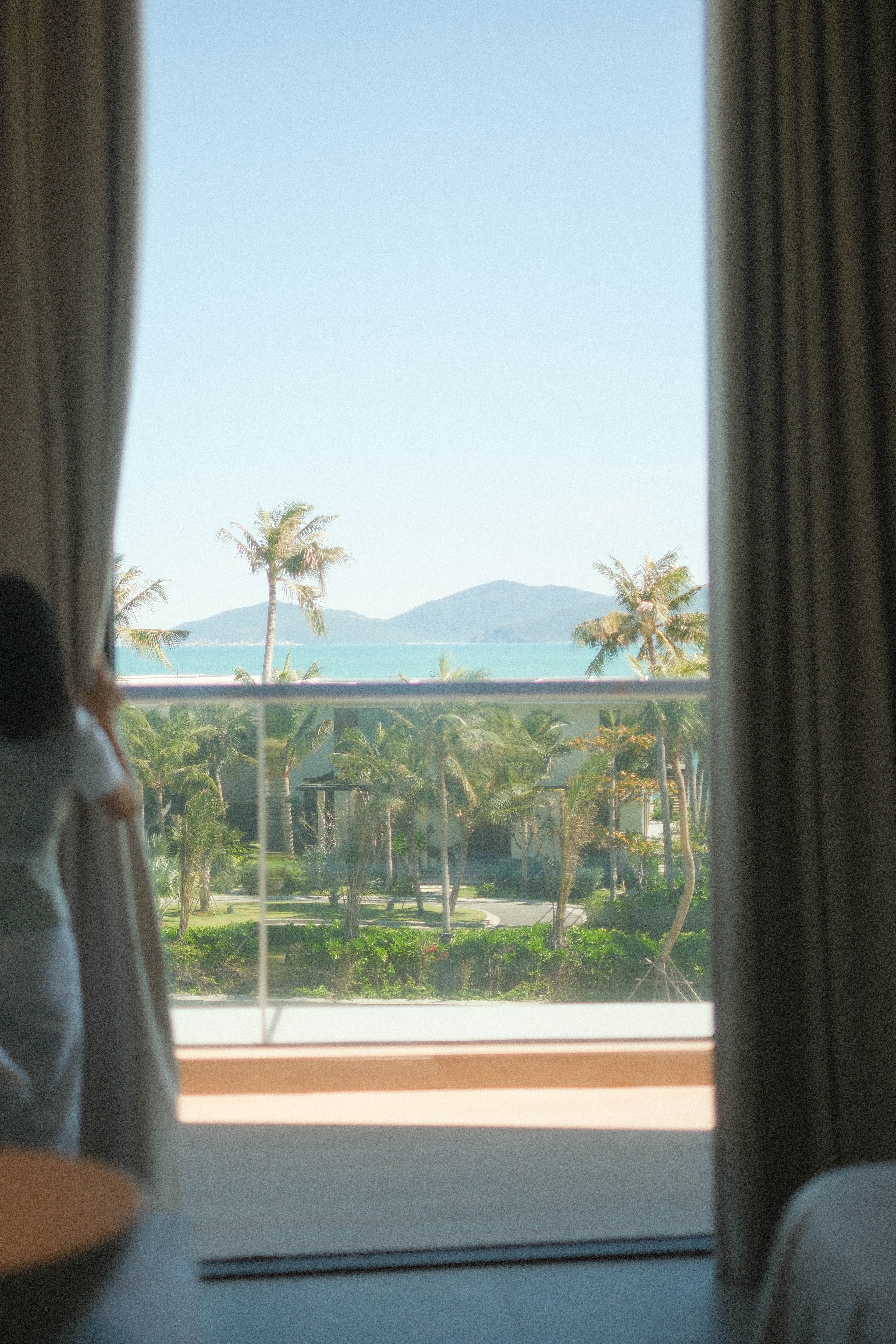 Tropical resort balcony view with ocean, palm trees, and mountains.