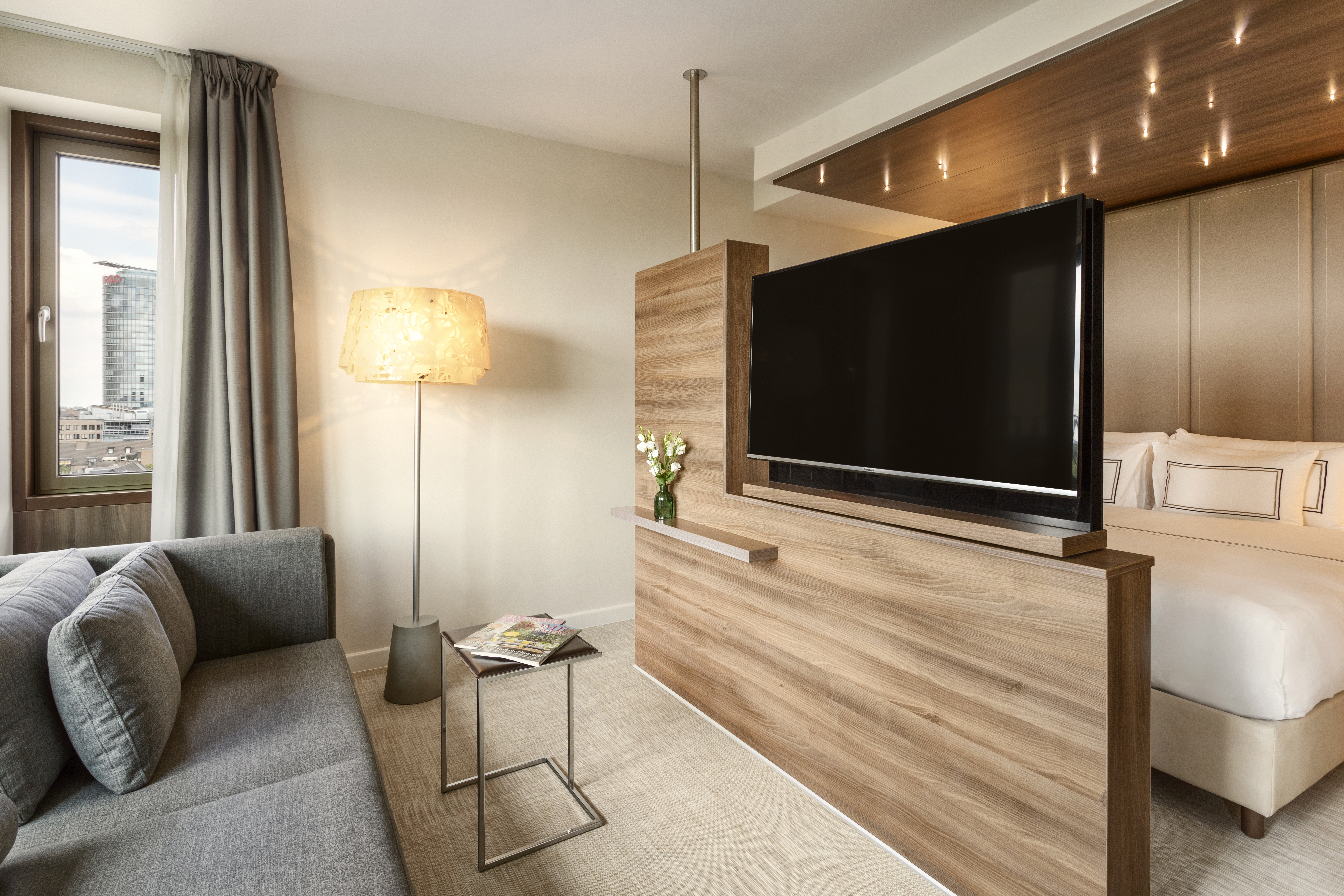 Modern hotel room with living area, flat-screen TV, sofa, bed, and city view.