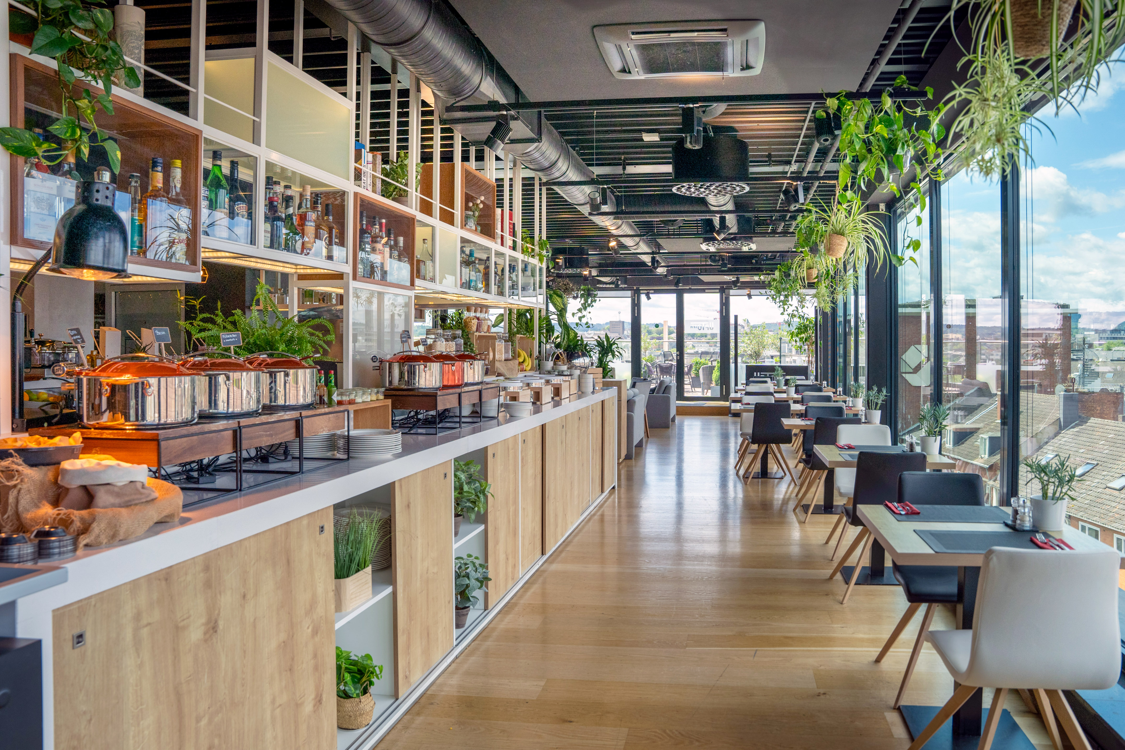 Modern restaurant buffet with food warmers, bar, dining tables, and city view.