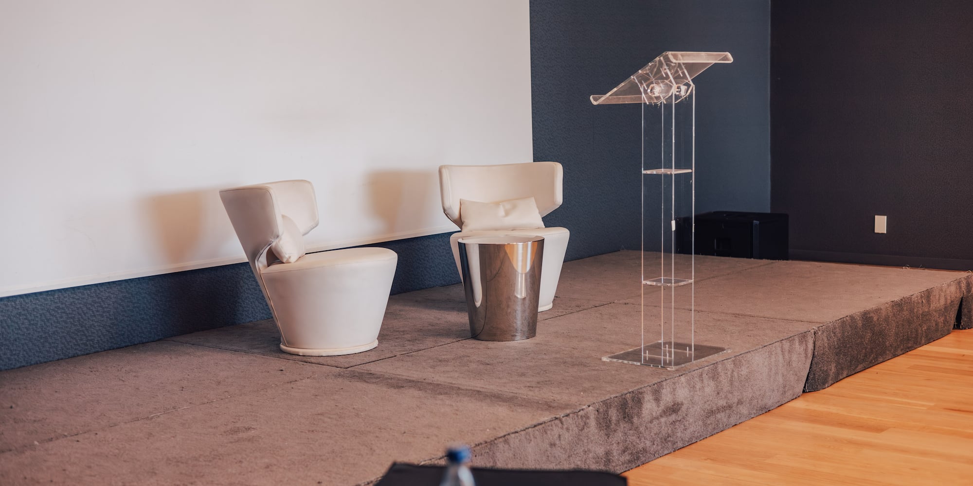 Empty conference stage with white armchairs, side table, and clear lectern.
