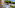 Balcony with white table, chairs, blue railing, street view, and large pine tree.