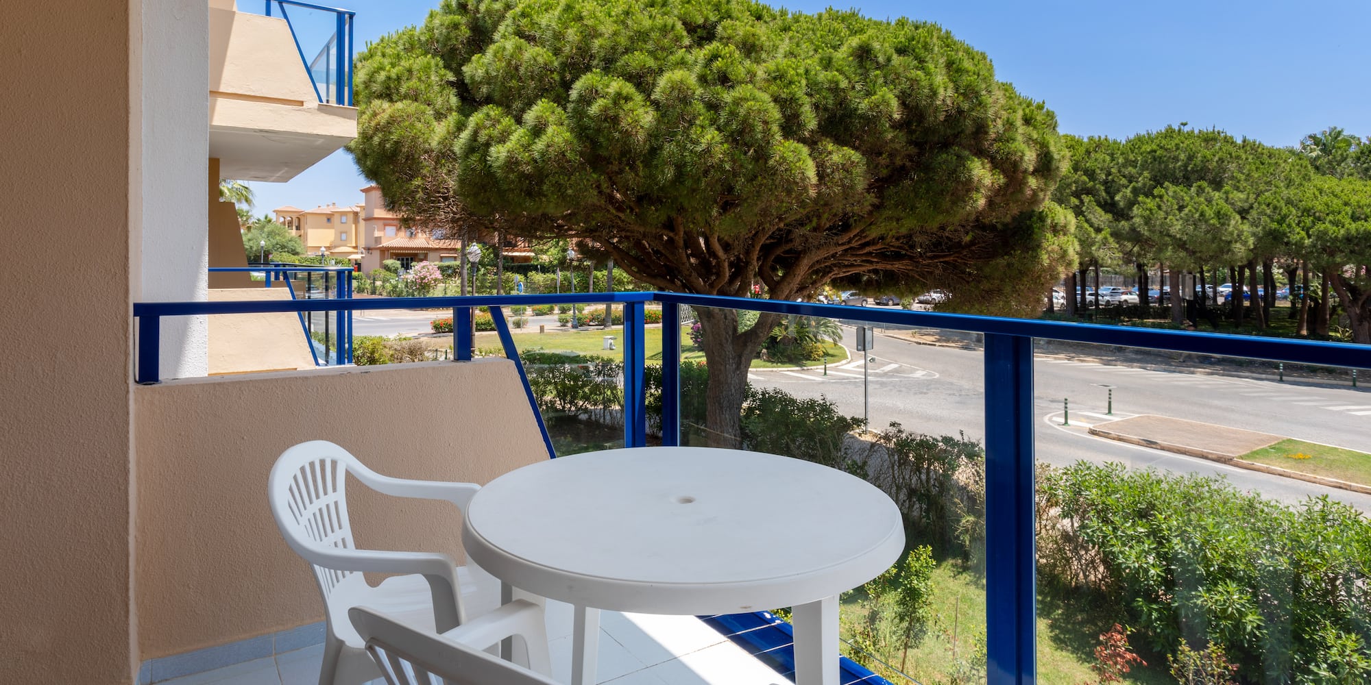 Balcony with white table, chairs, blue railing, street view, and large pine tree.