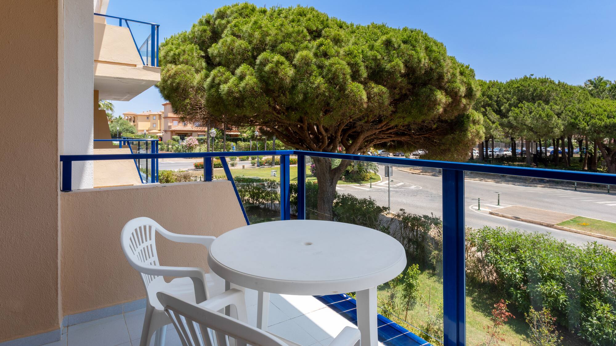Balcony with white table, chairs, blue railing, street view, and large pine tree.