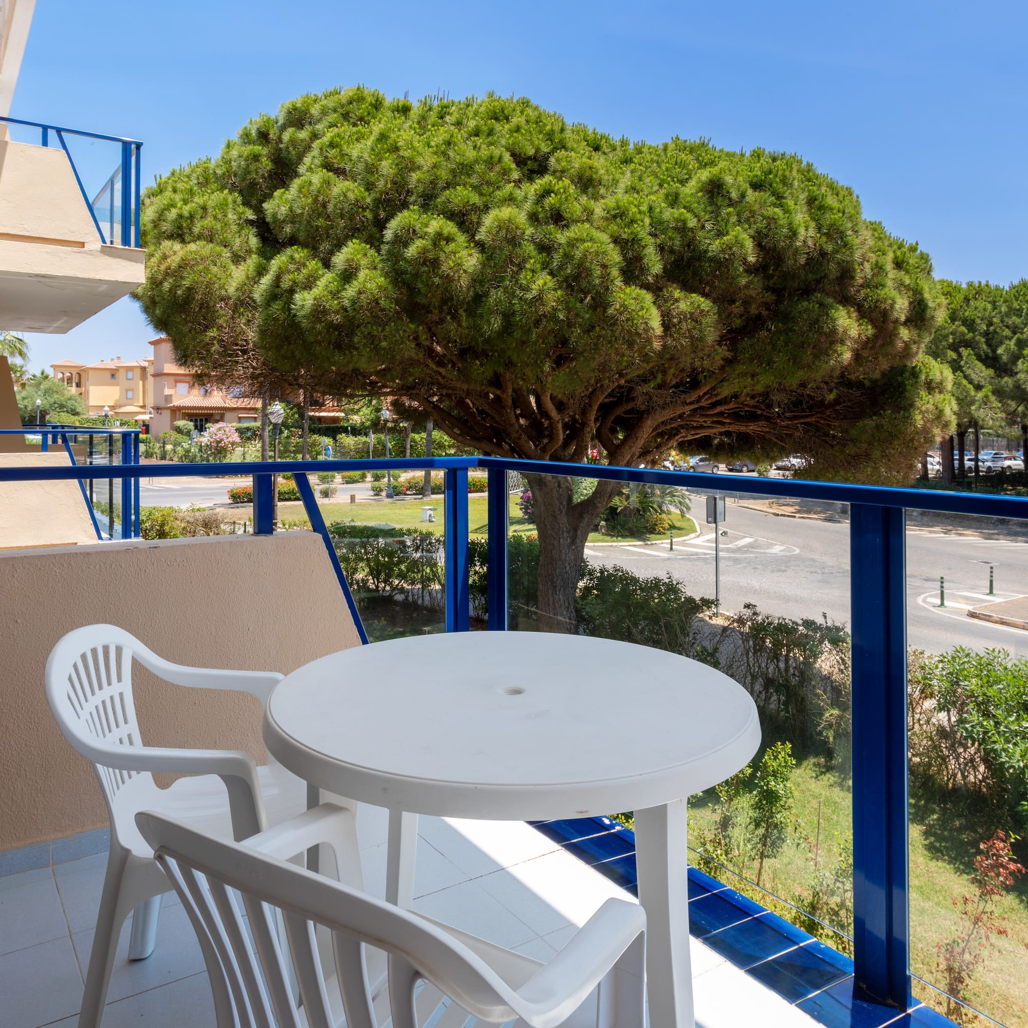 Balcony with white table, chairs, blue railing, street view, and large pine tree.