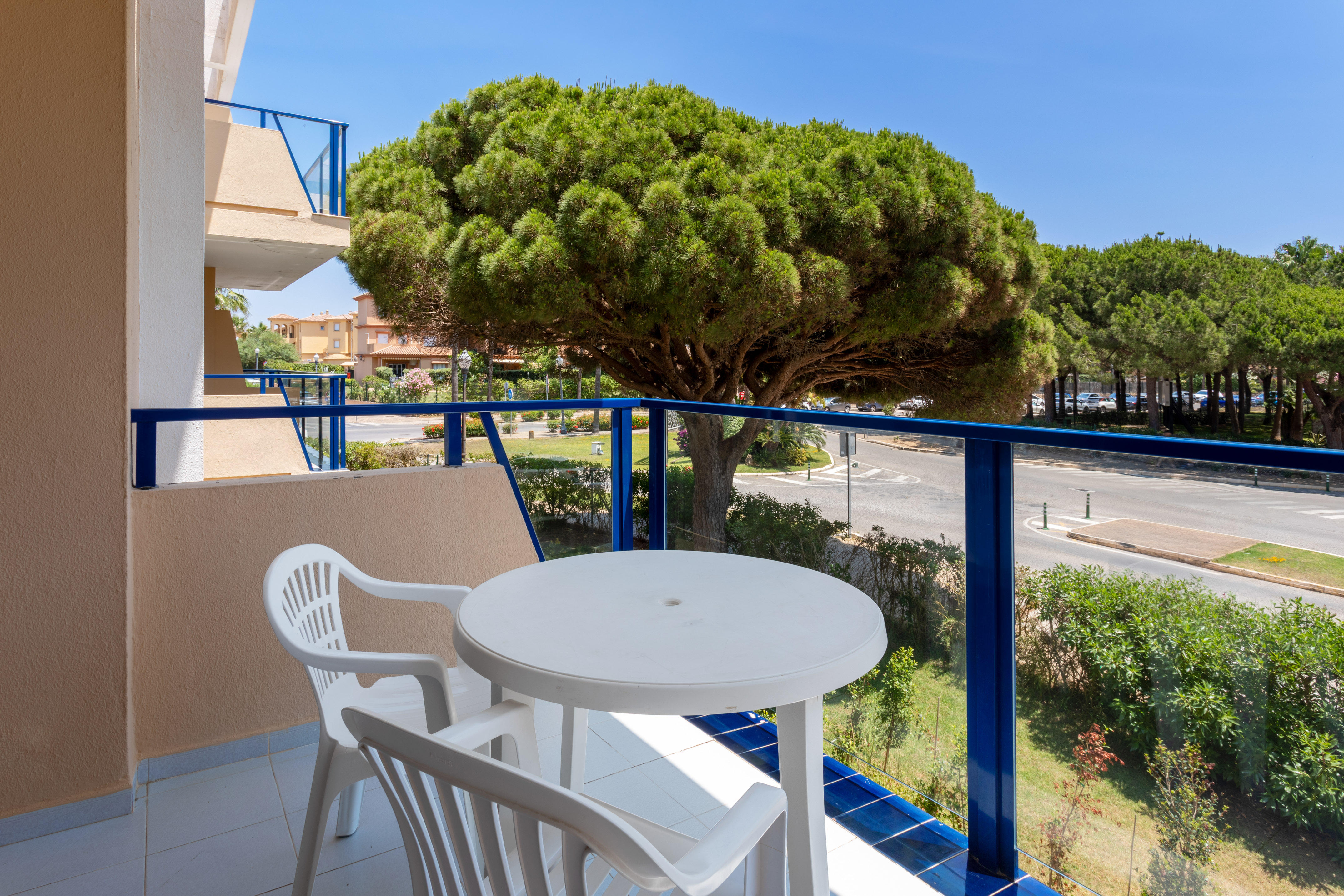 Balcony with white table, chairs, blue railing, street view, and large pine tree.