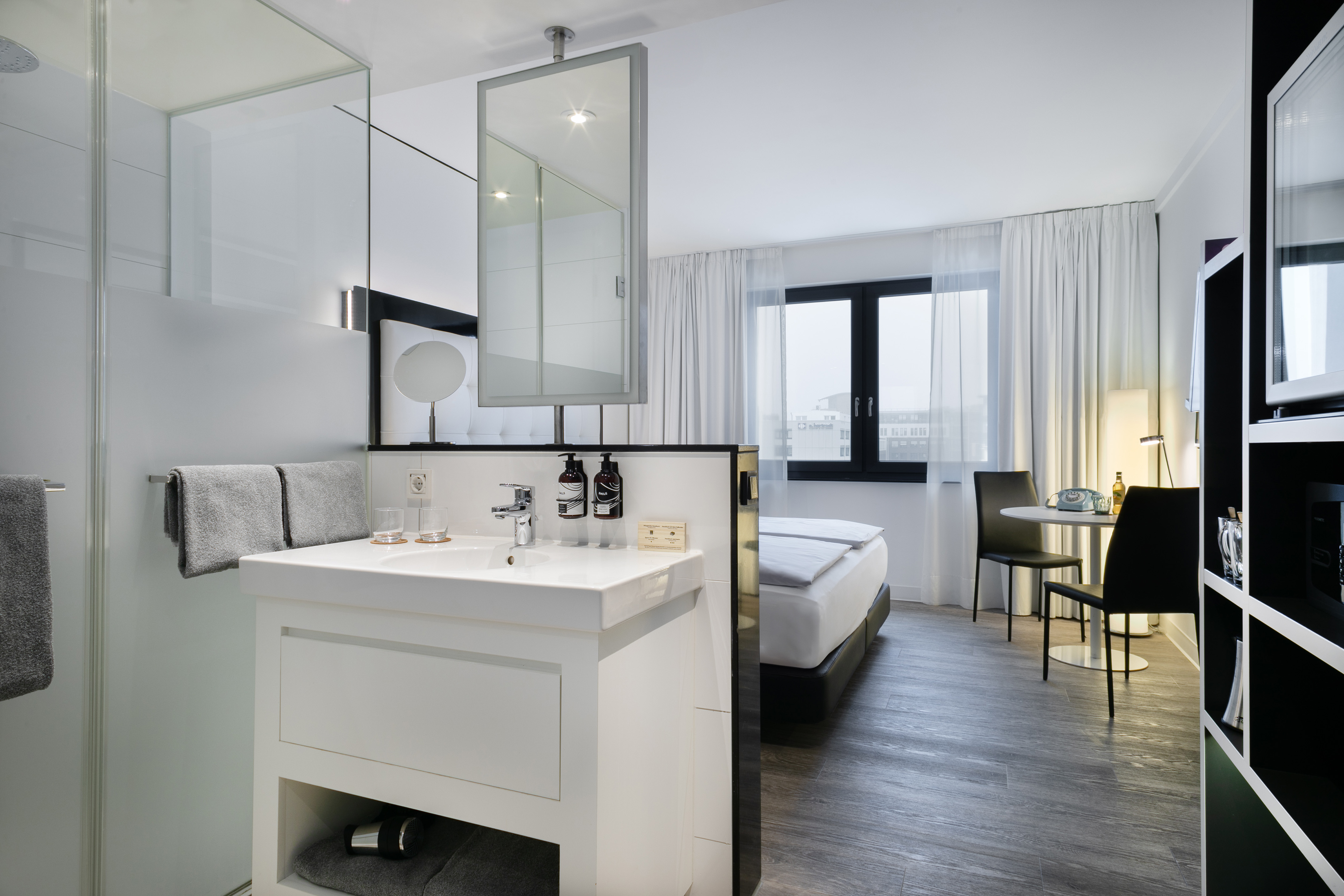 Modern studio hotel room with integrated bathroom sink, bed, and small desk.