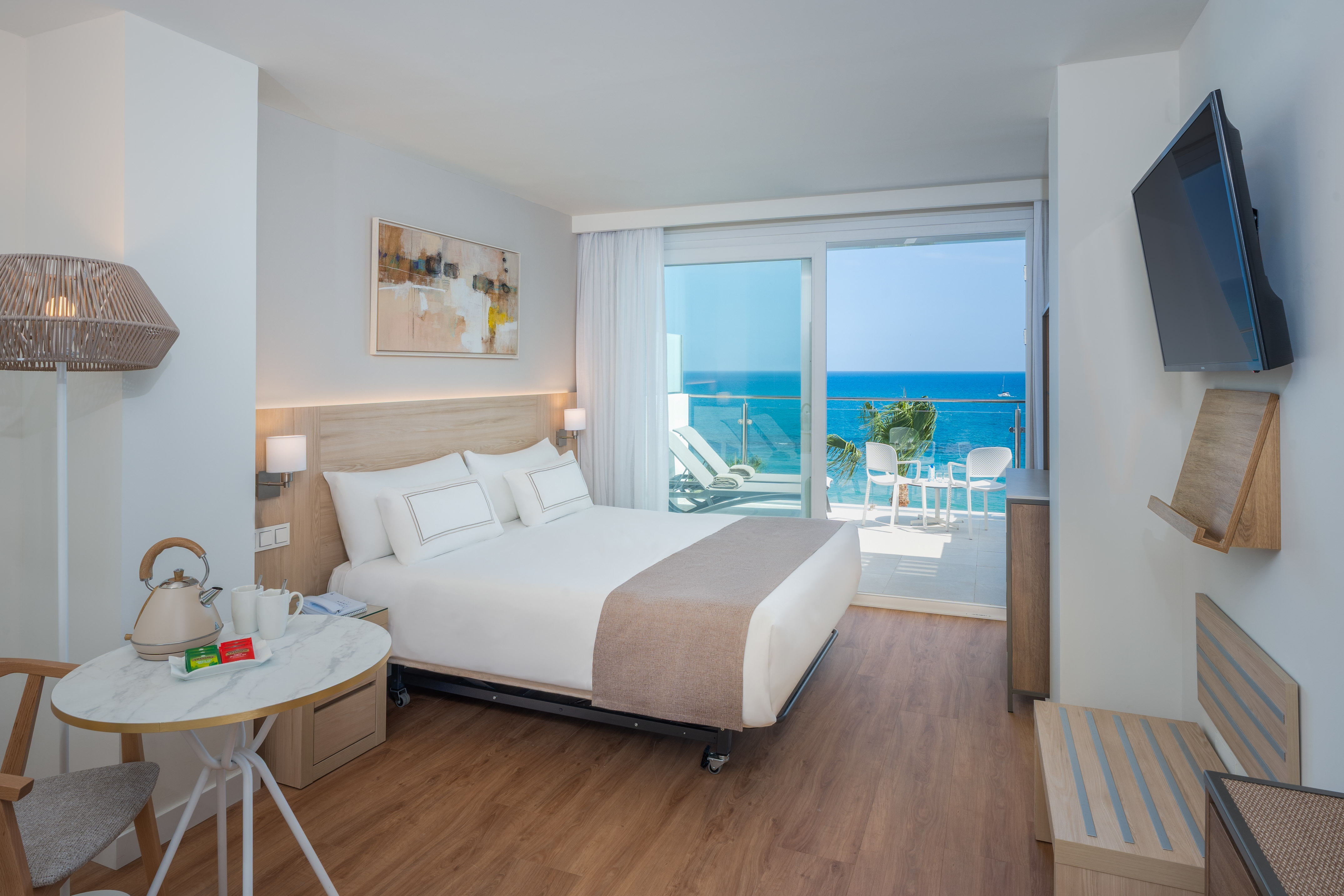 Modern hotel room with ocean view balcony, white bed, and wooden floor.