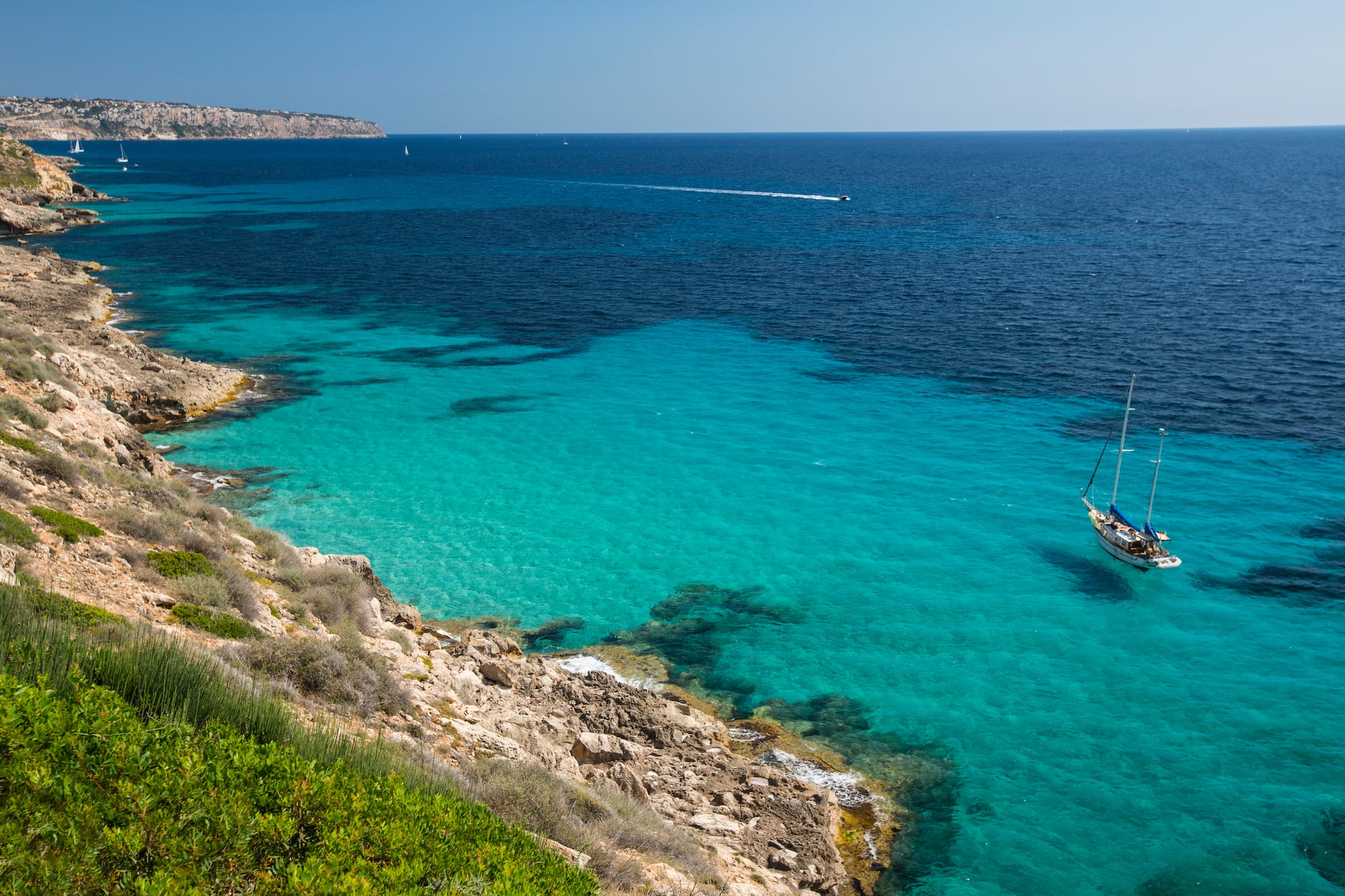 Velero en cala mediterránea de aguas turquesas y acantilados rocosos.