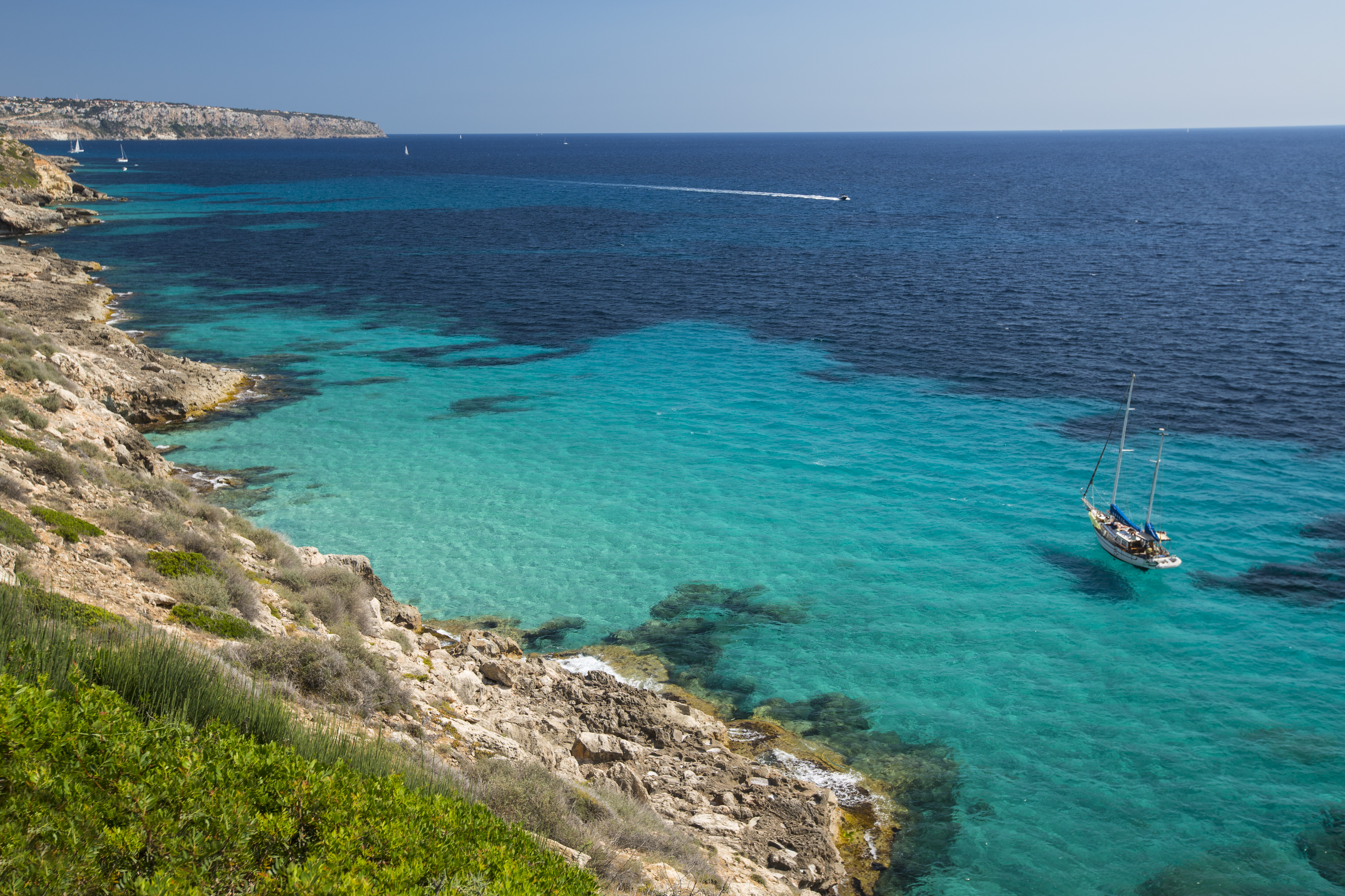Velero en cala mediterránea de aguas turquesas y acantilados rocosos.