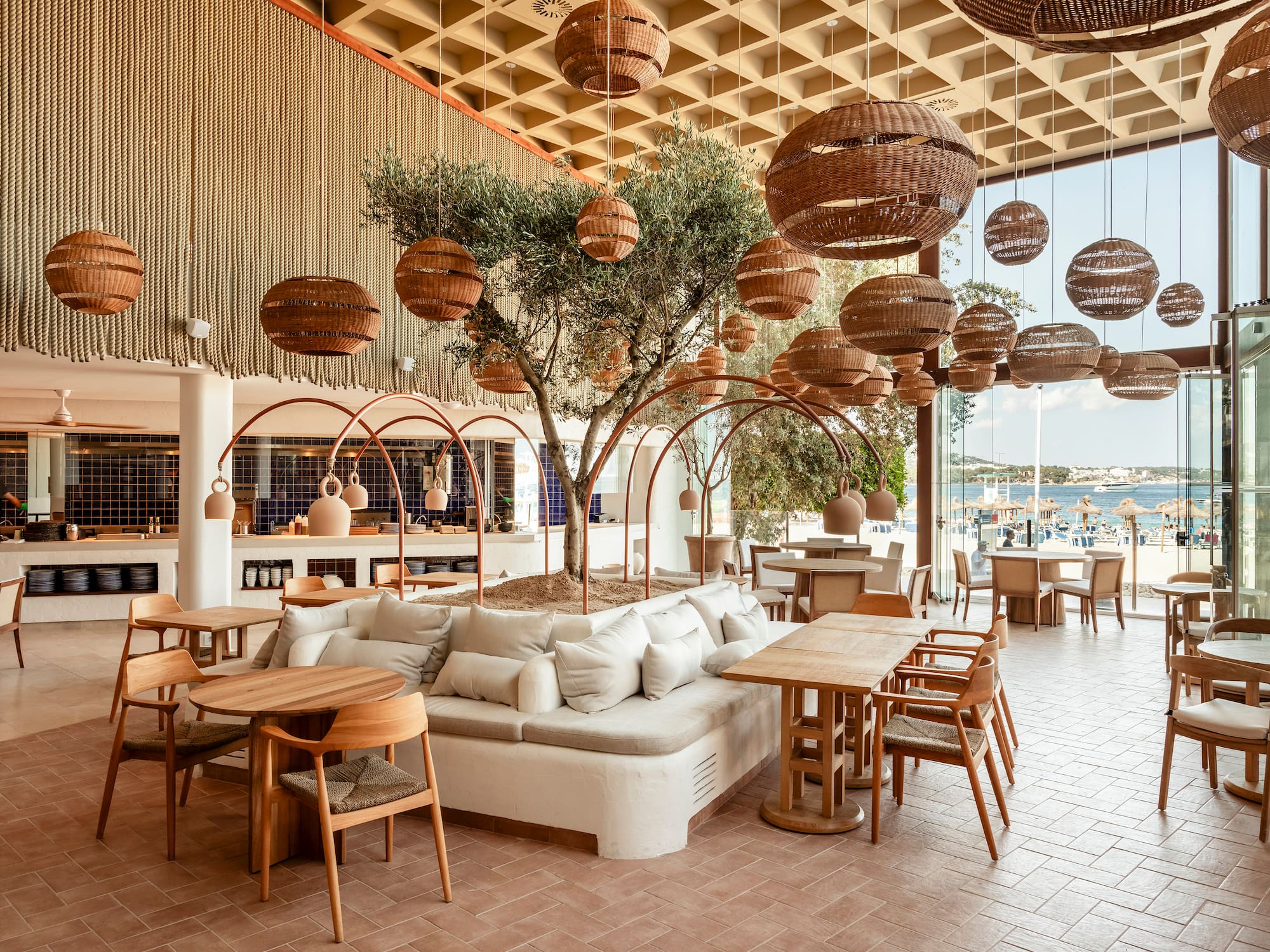 Modern restaurant interior with olive tree, woven lights, and beach view.