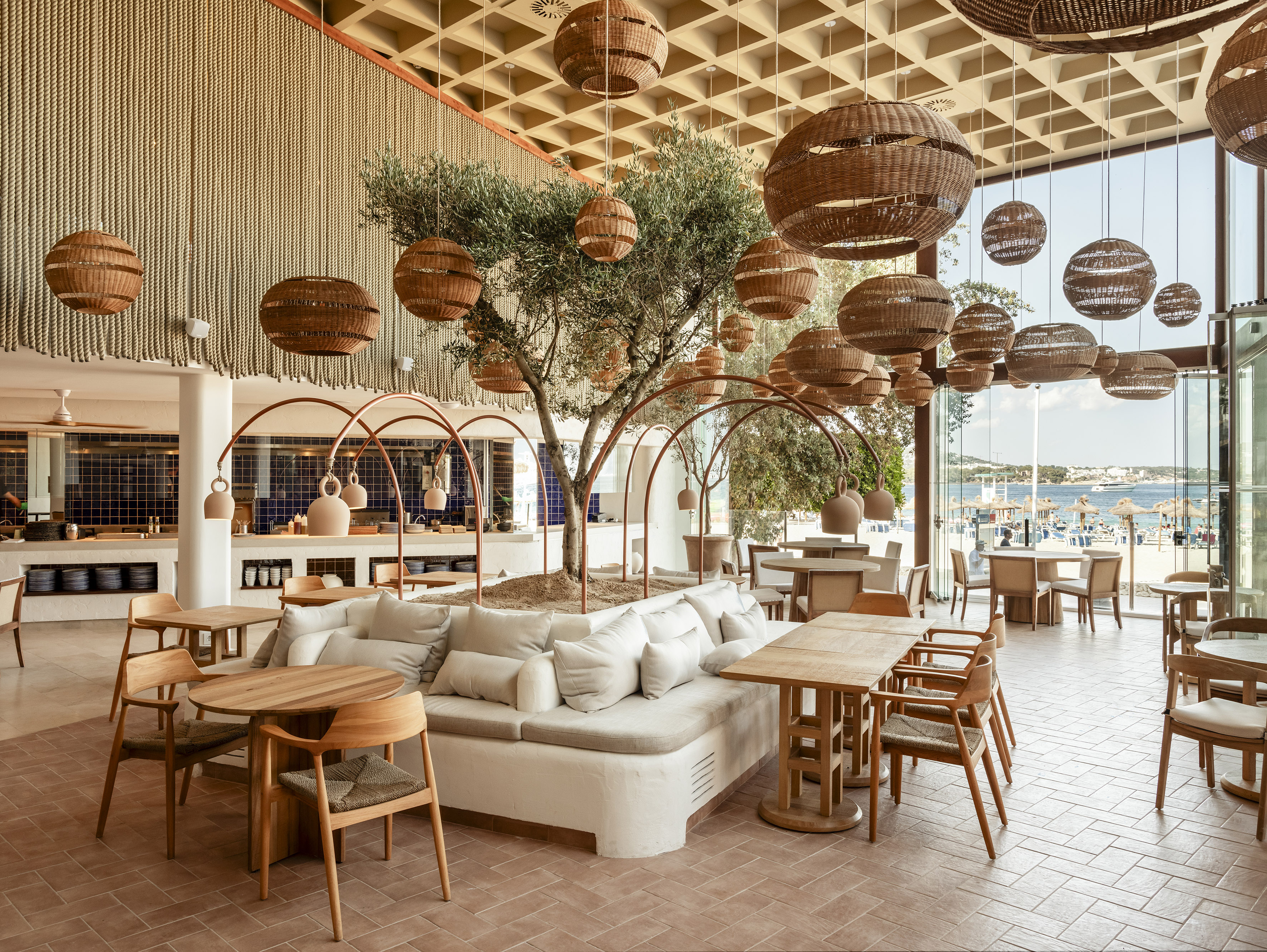 Modern restaurant interior with olive tree, woven lights, and beach view.