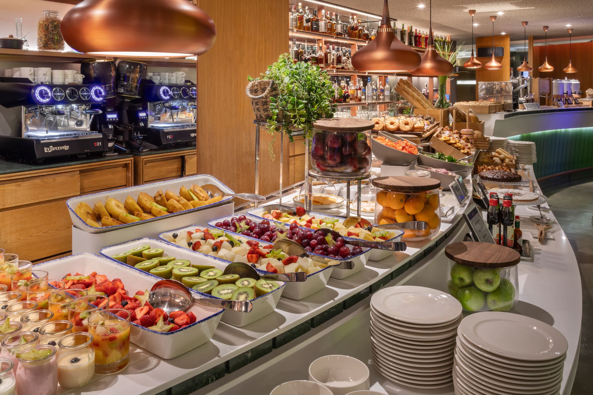 Hotel breakfast buffet with fresh fruit, pastries, coffee machines, and desserts.