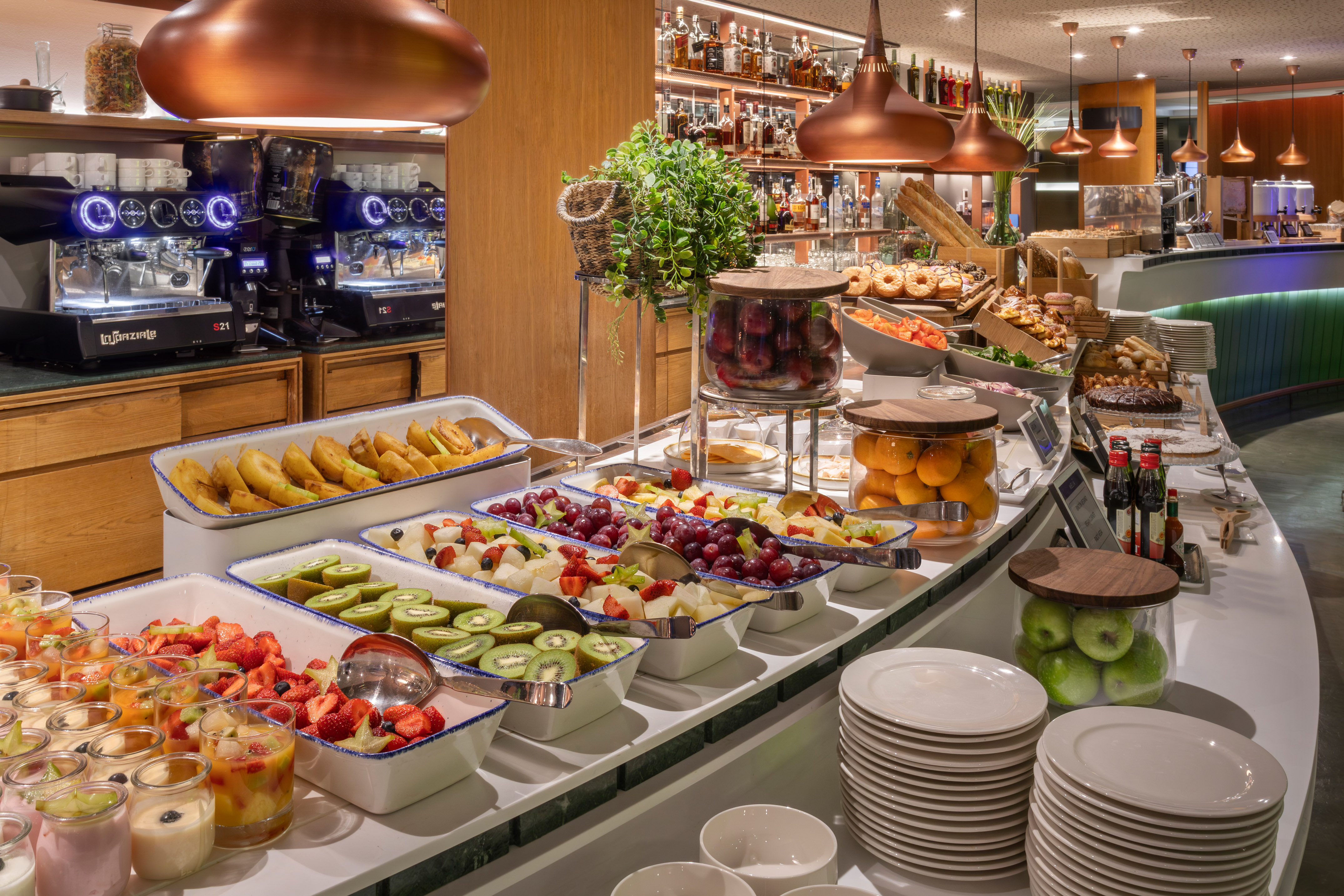 Hotel breakfast buffet with fresh fruit, pastries, coffee machines, and desserts.