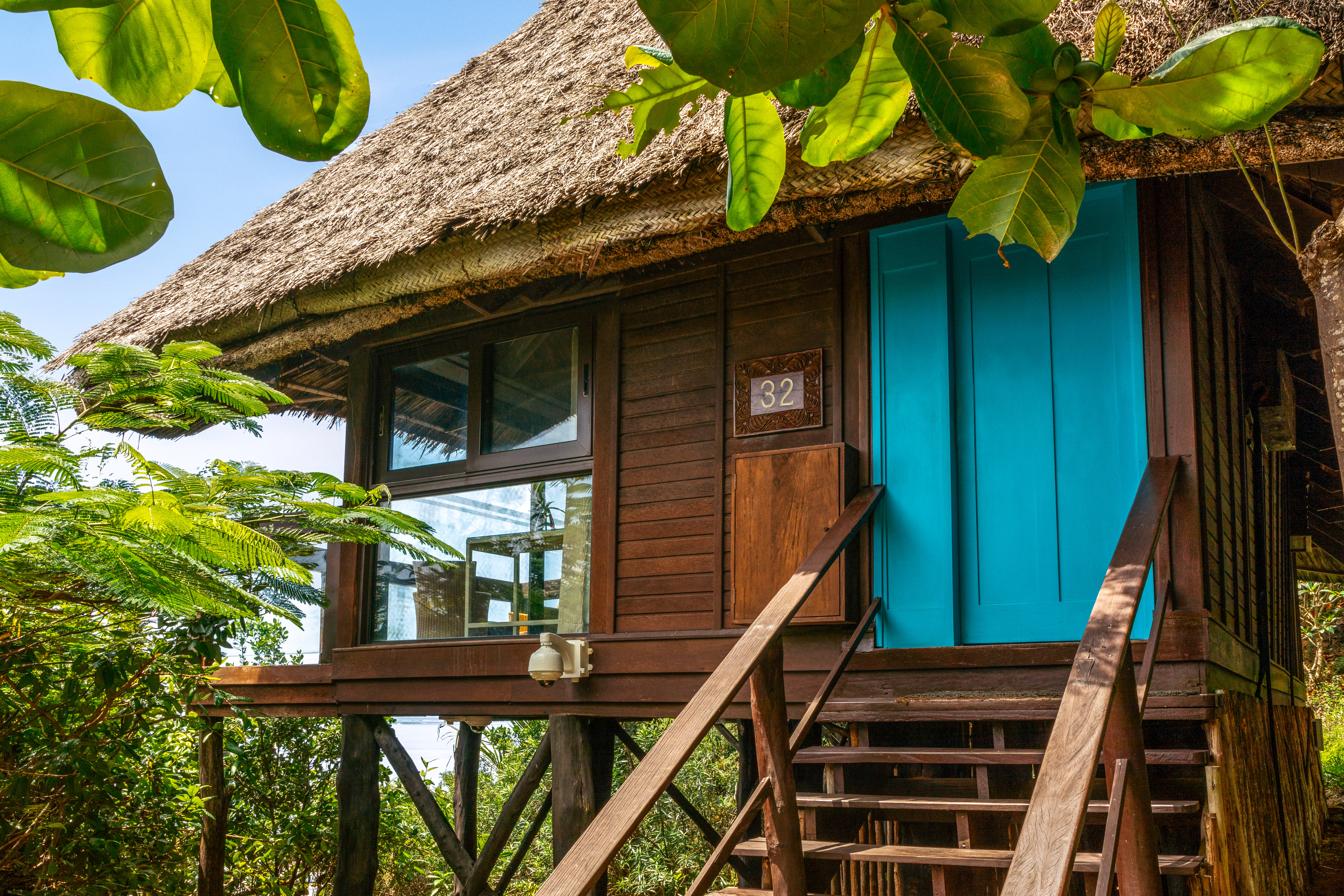 Tropical stilt house #32 with thatched roof, vibrant blue door, and wooden steps.