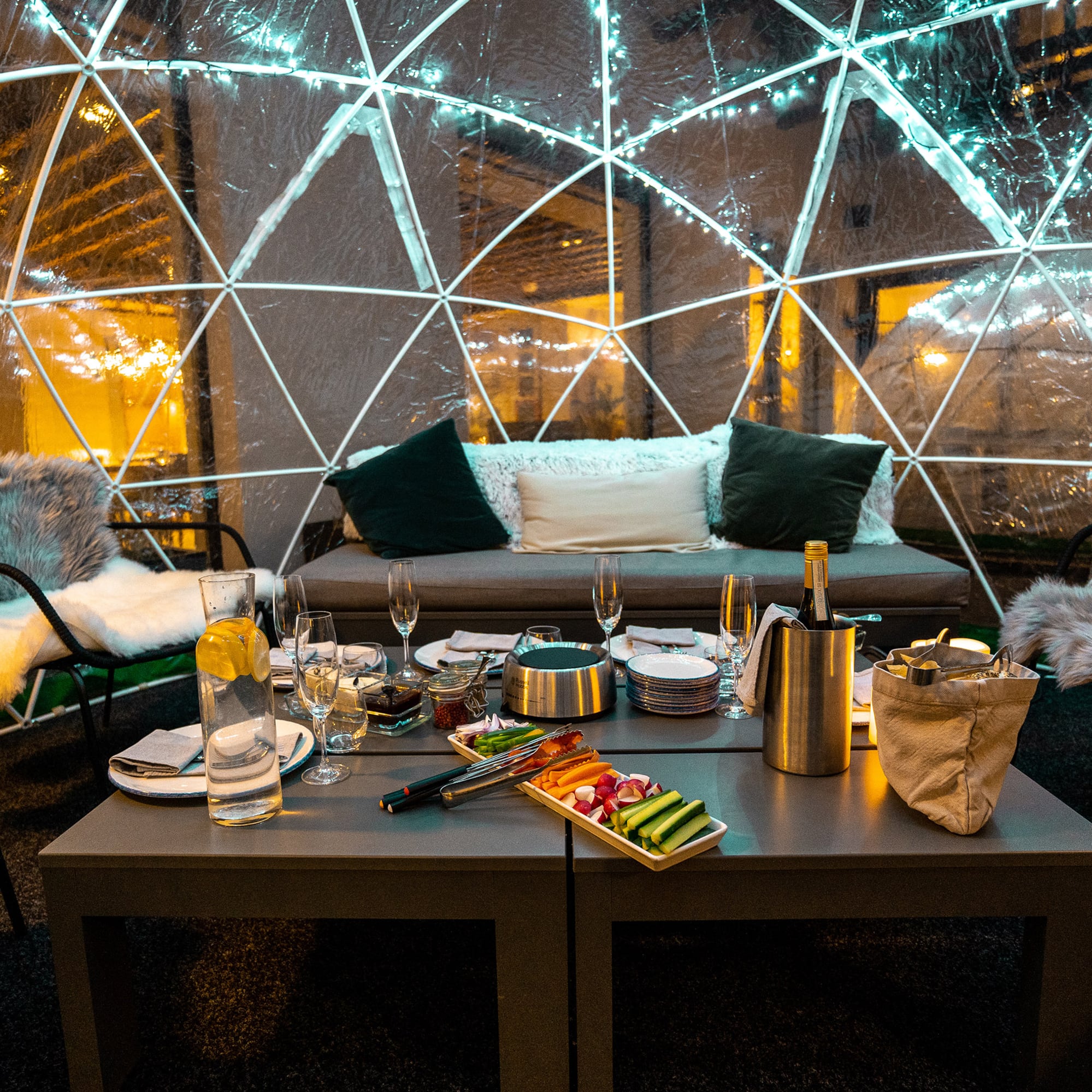 Cozy igloo dining setup with food, champagne, and string lights.