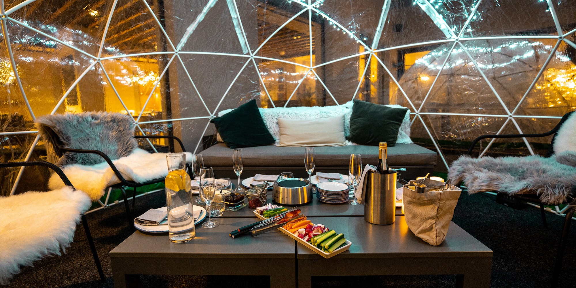 Cozy igloo dining setup with food, champagne, and string lights.