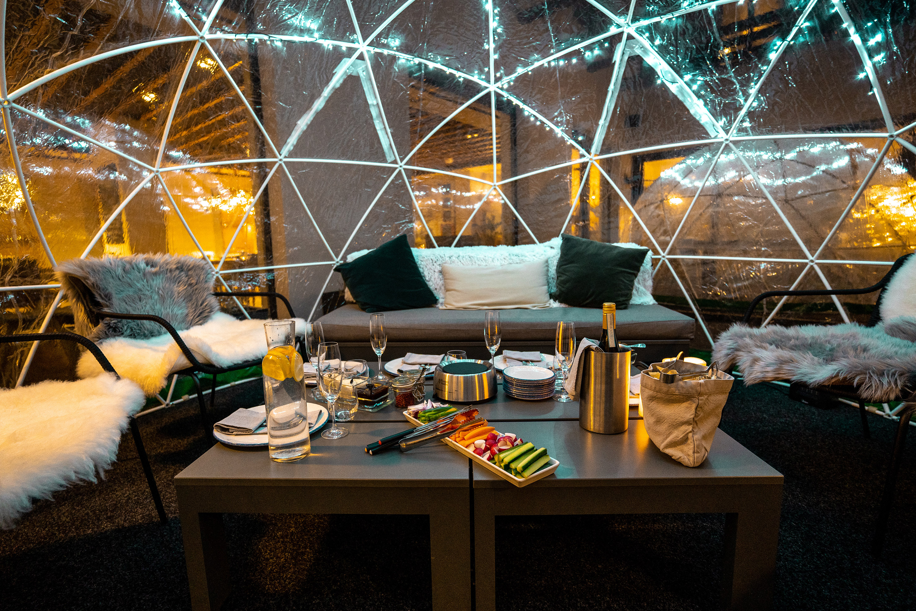 Cozy igloo dining setup with food, champagne, and string lights.