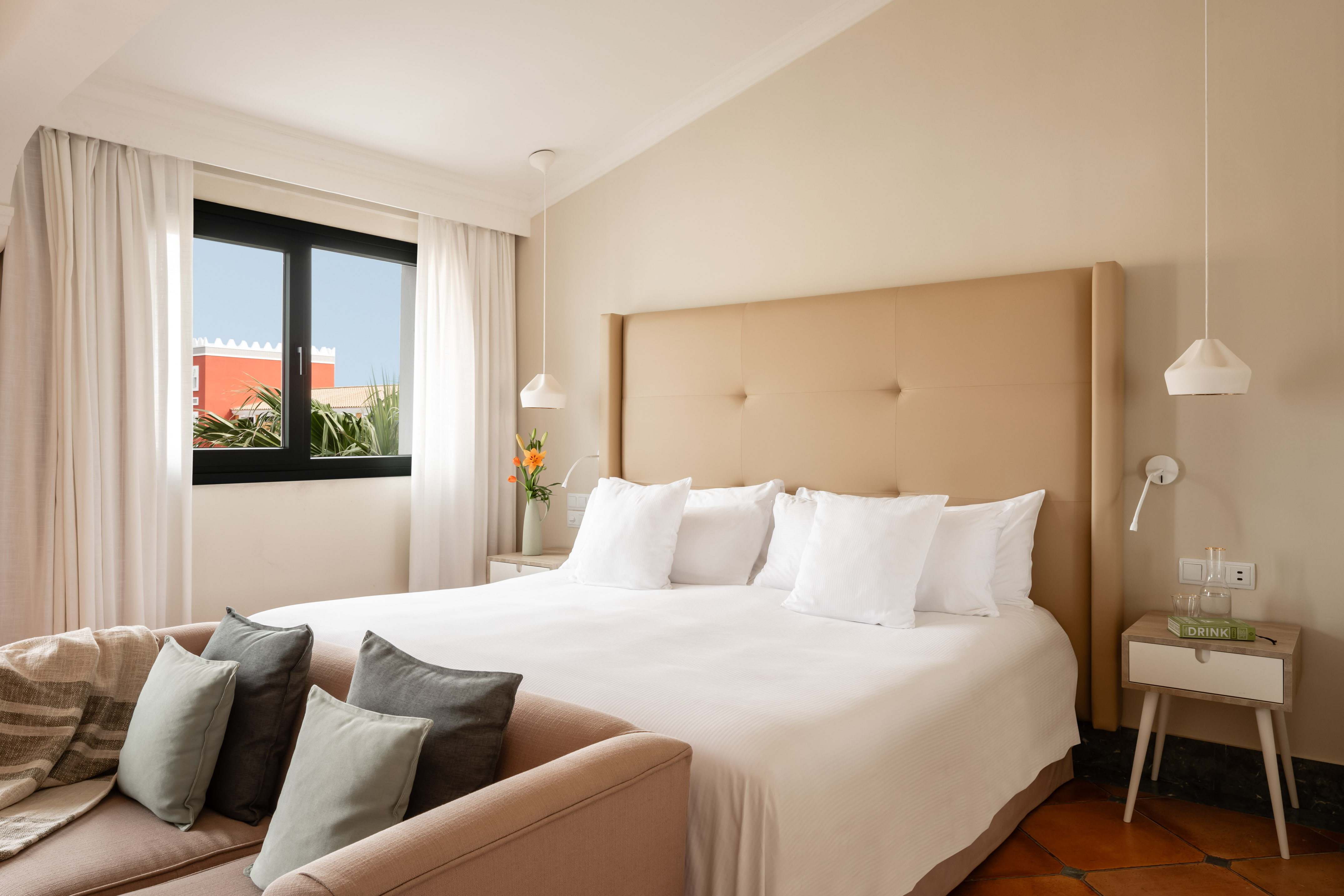 Modern bedroom with large bed, beige headboard, window, and lounge seating.