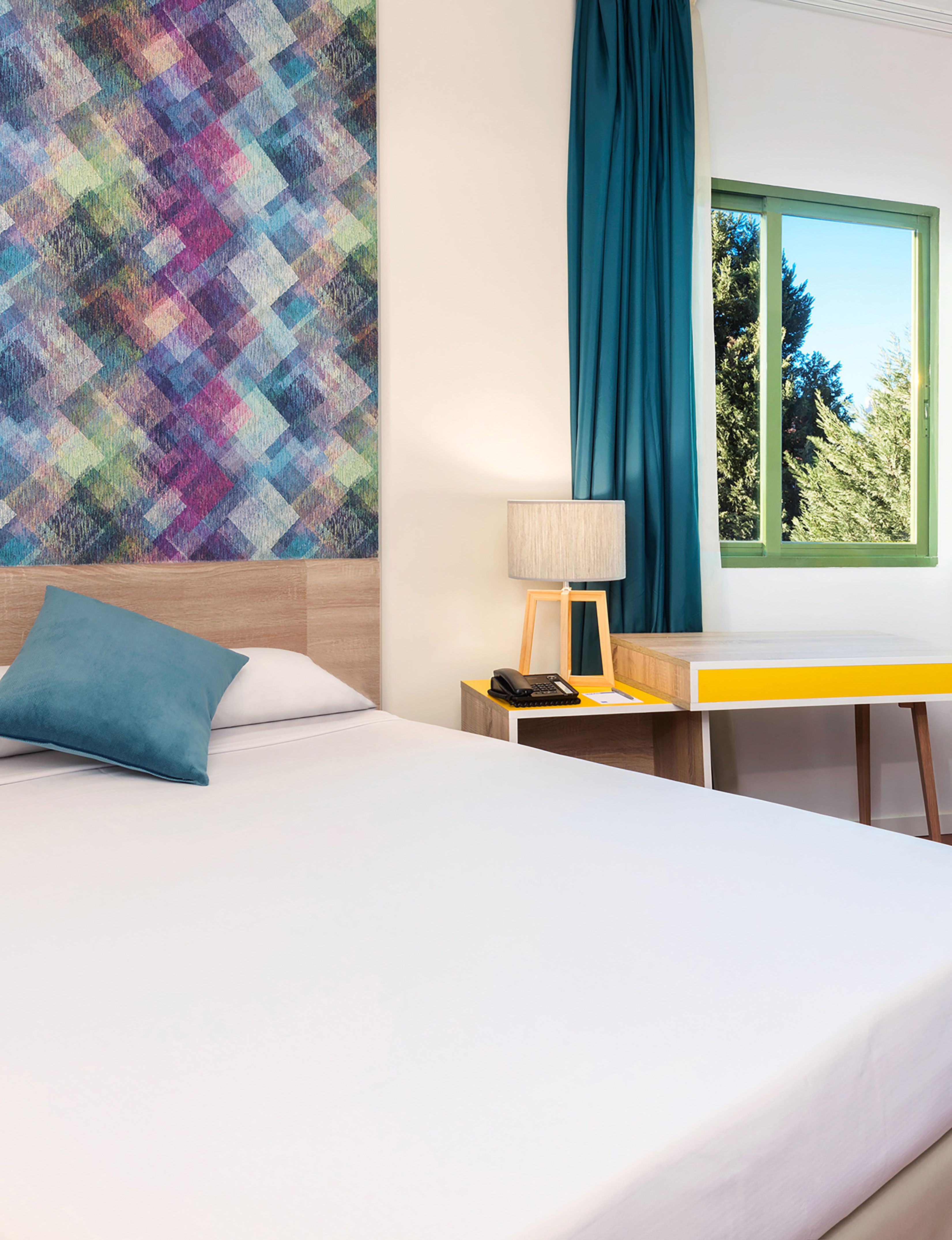 Modern hotel room with white bed, colorful geometric art, window, and bedside desk.