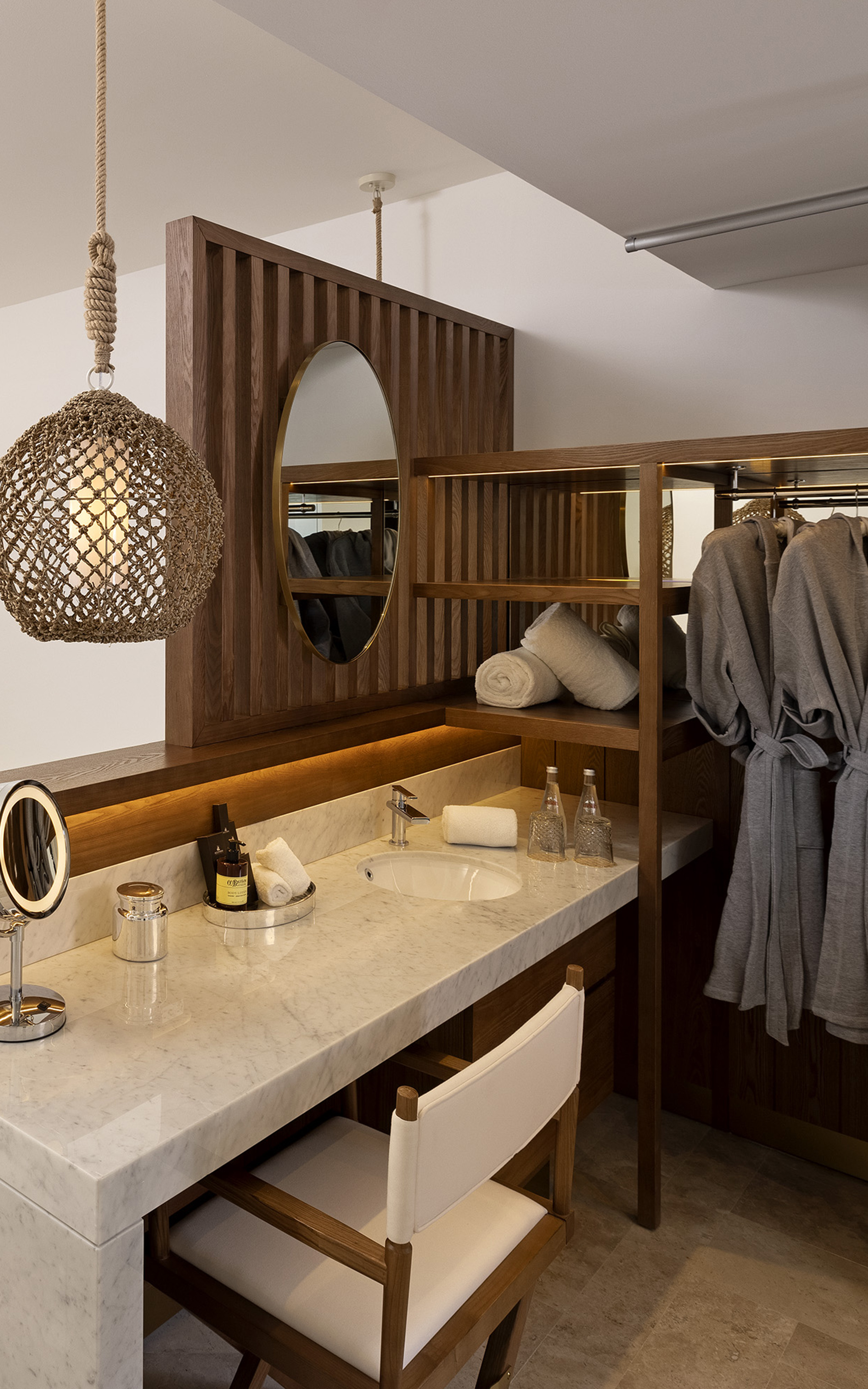 Marble bathroom vanity, wooden shelving, sink, robes, and rope pendant light.