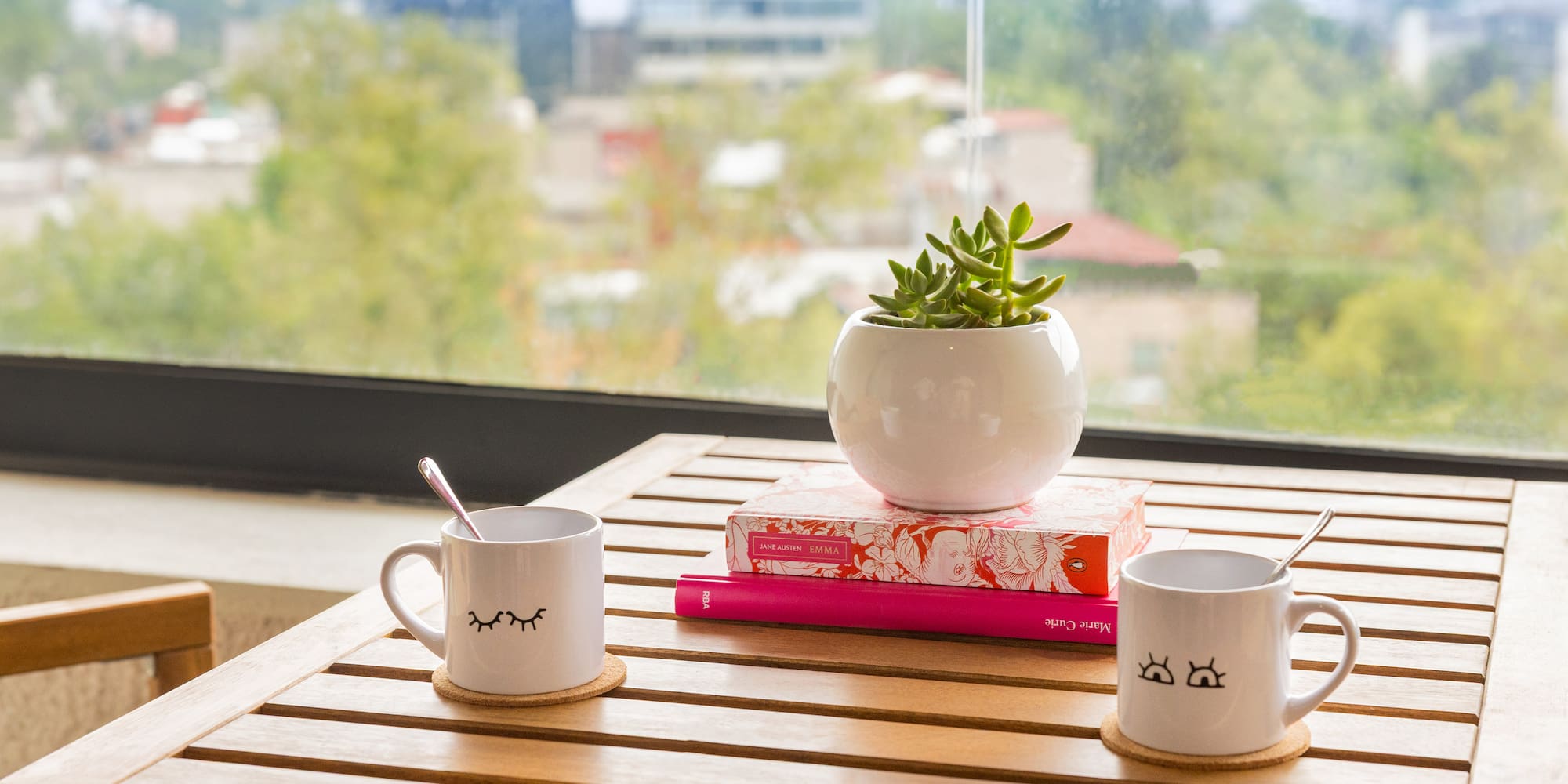 Wooden table with two eye mugs, succulent, books, and city view.