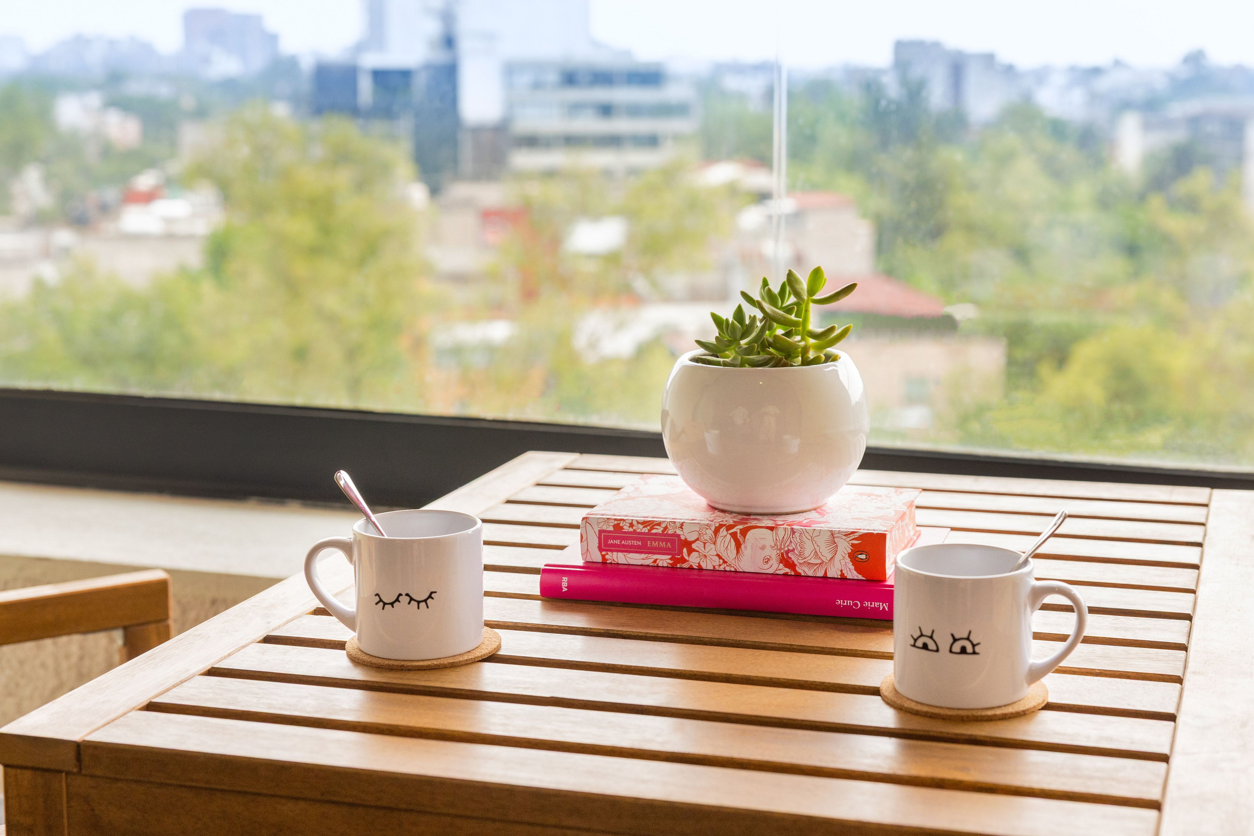 Two eye-design mugs, succulent, books on wooden table, city balcony view.