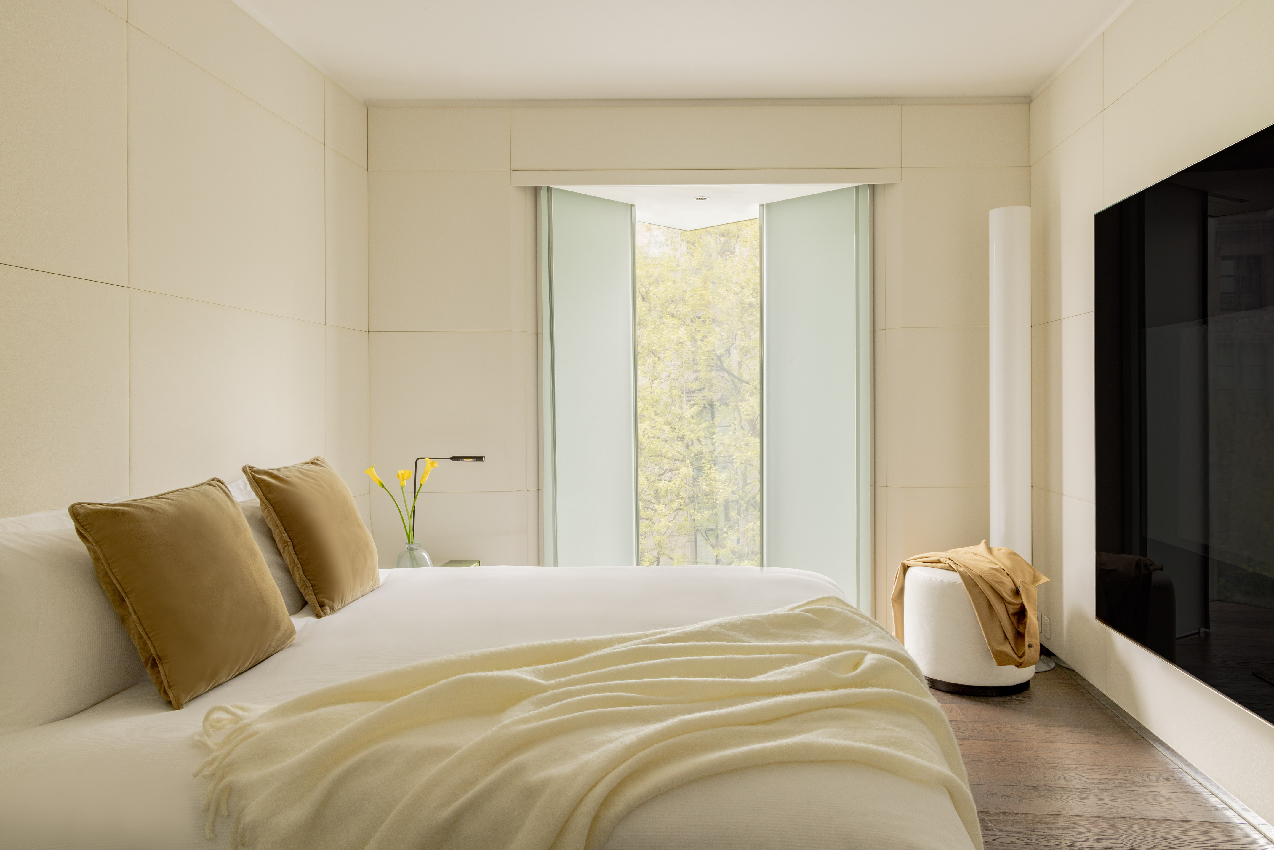 Modern hotel room with white bed, gold pillows, large window, and wood floor.