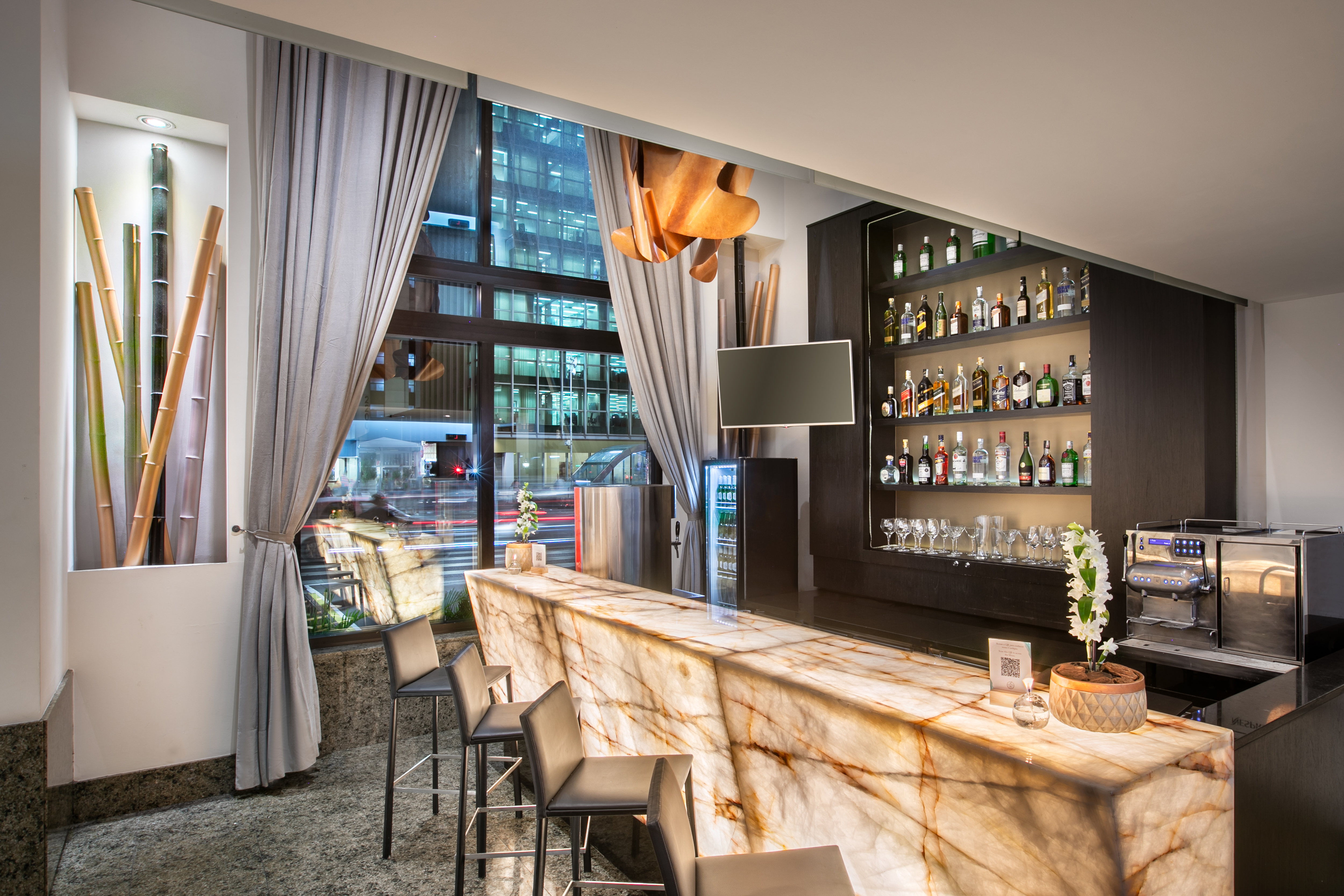 Hotel bar with illuminated marble counter, liquor display, and cityscape view.