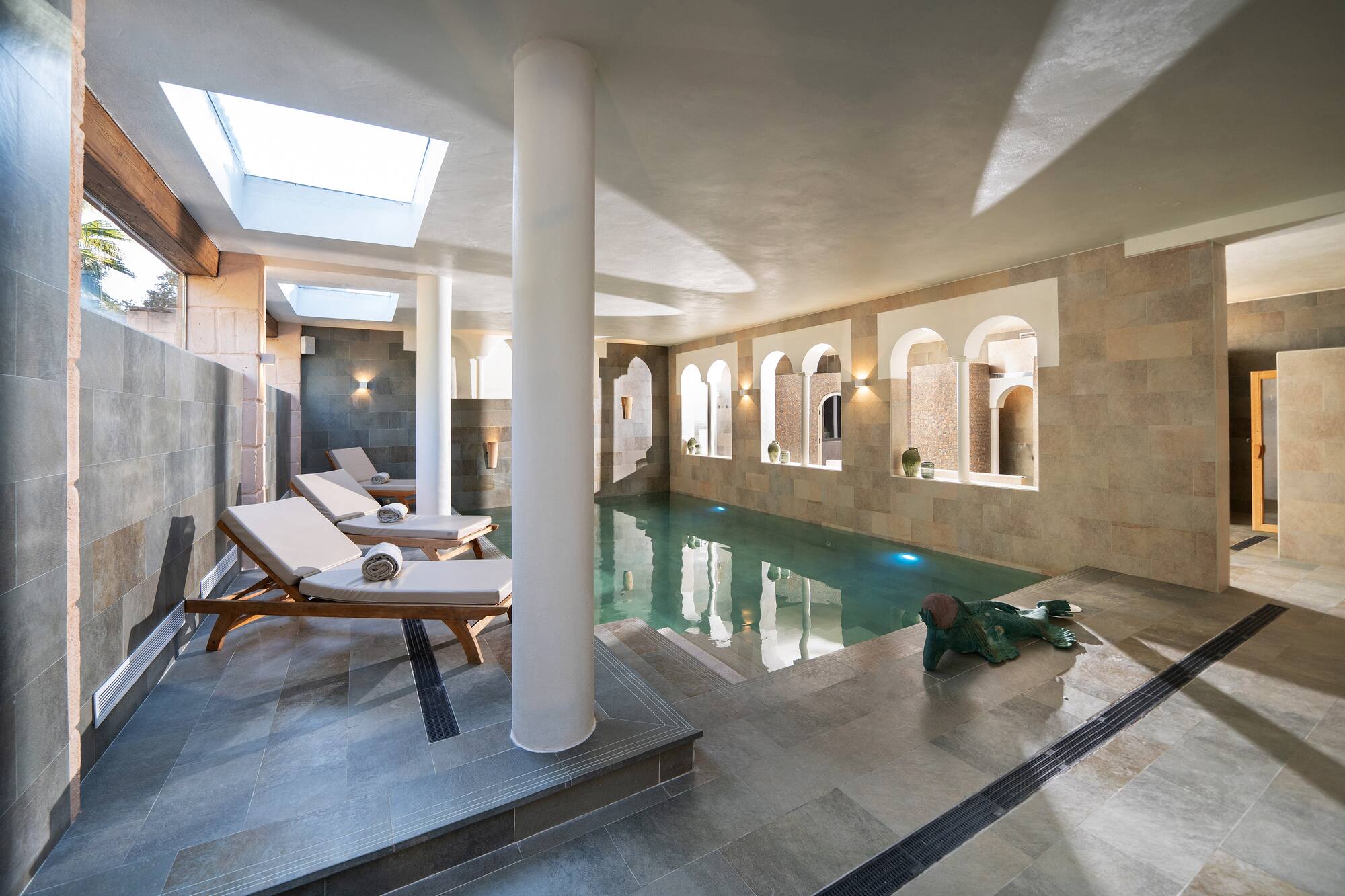 Elegant indoor spa pool with skylights, stone tiles, and lounge chairs.