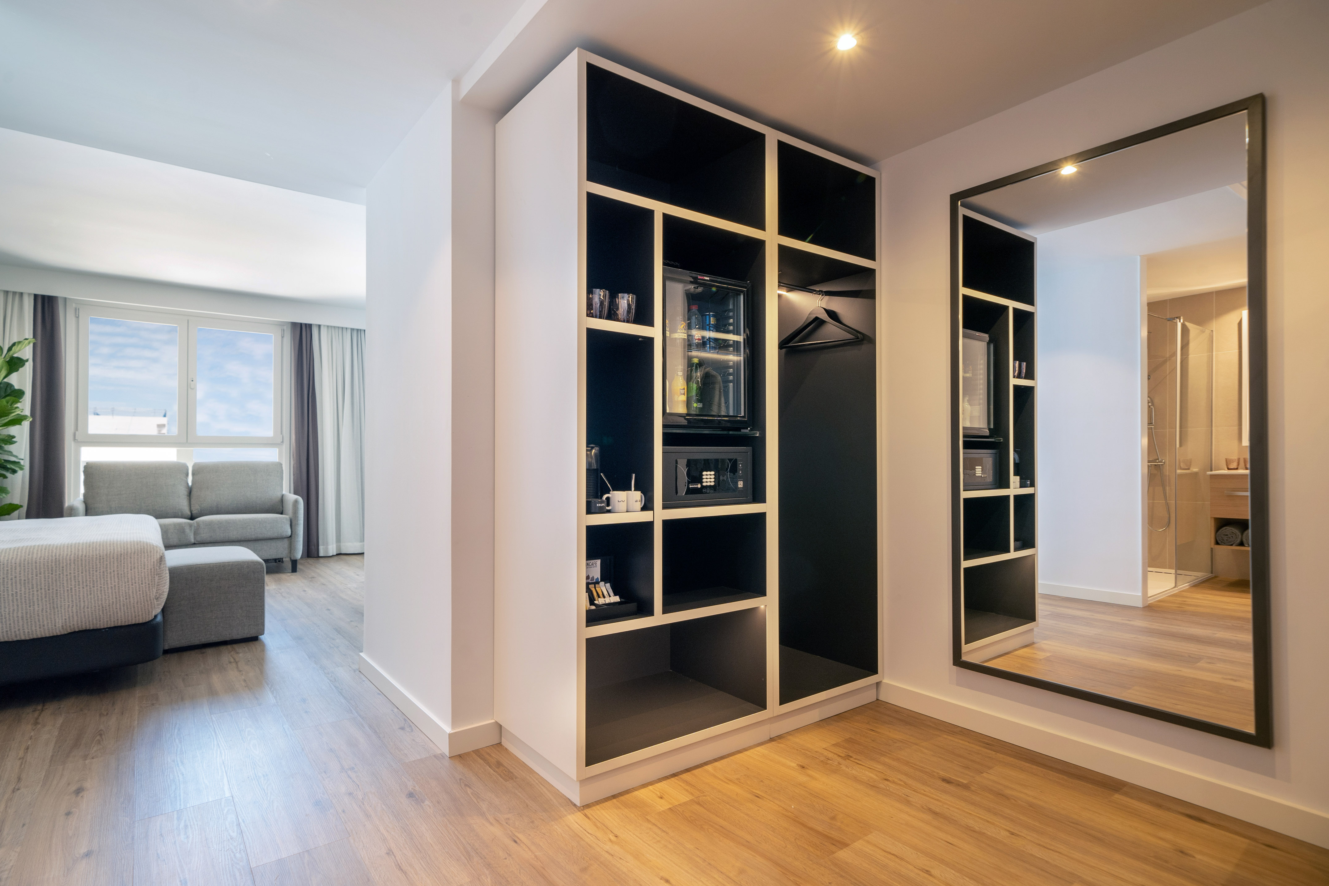 Modern hotel suite with wardrobe, minibar, safe, and mirror reflecting bathroom.