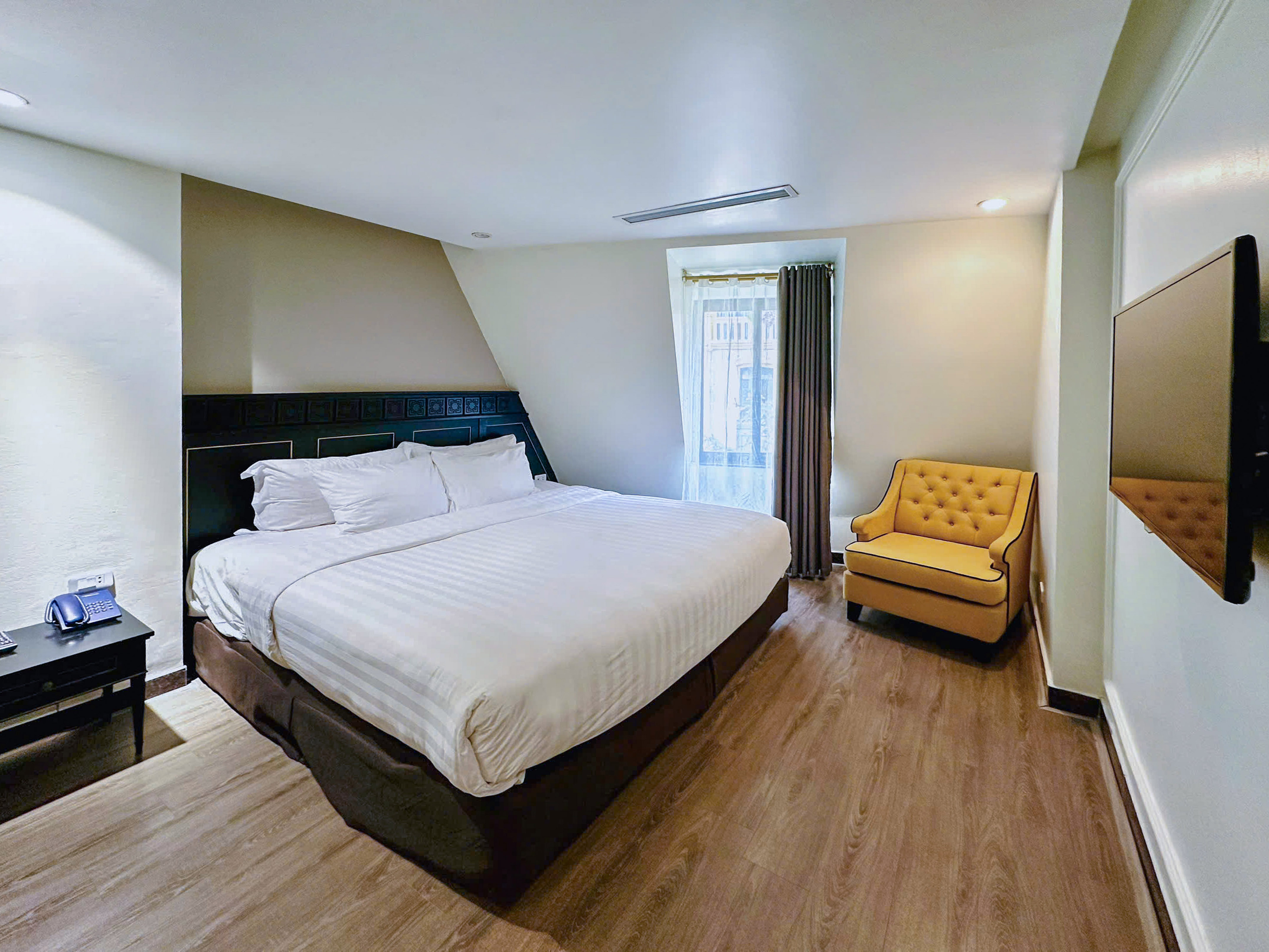Hotel bedroom with large white bed, yellow accent chair, wood floor, and window.
