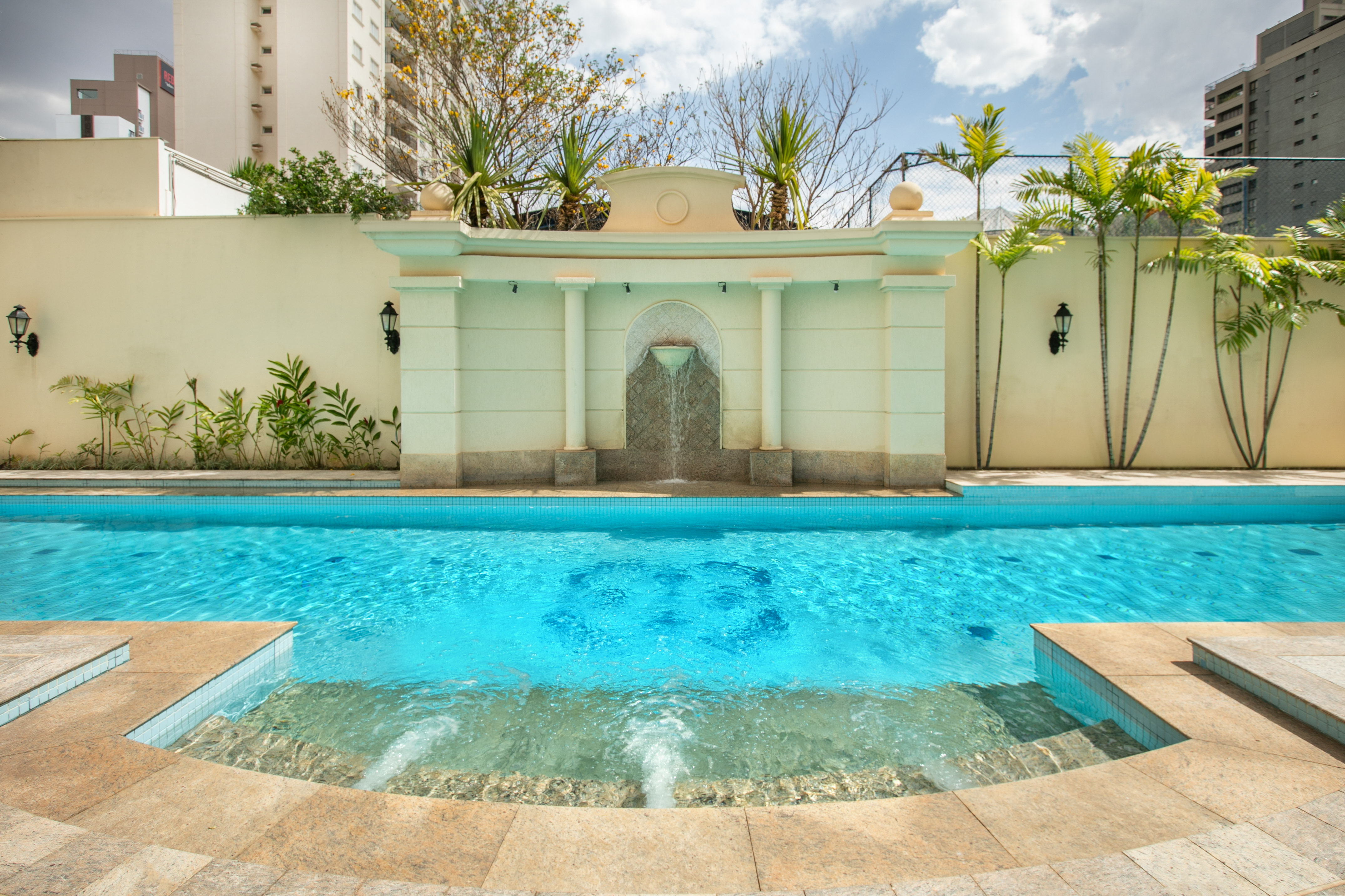 Luxury outdoor swimming pool with fountain, palm trees, and building backdrop.