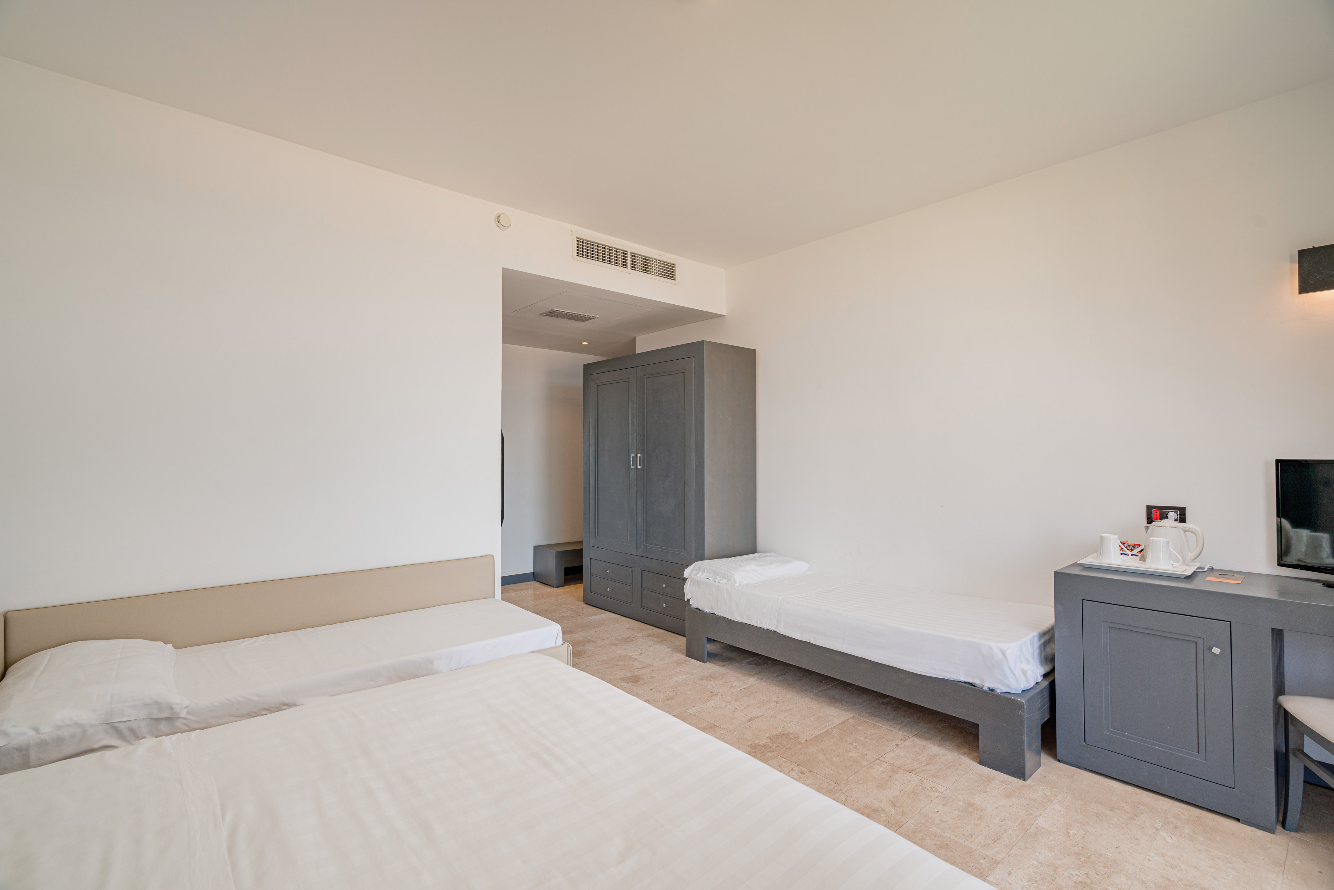 Clean hotel room with three beds, grey wardrobe, and light tiled floor.