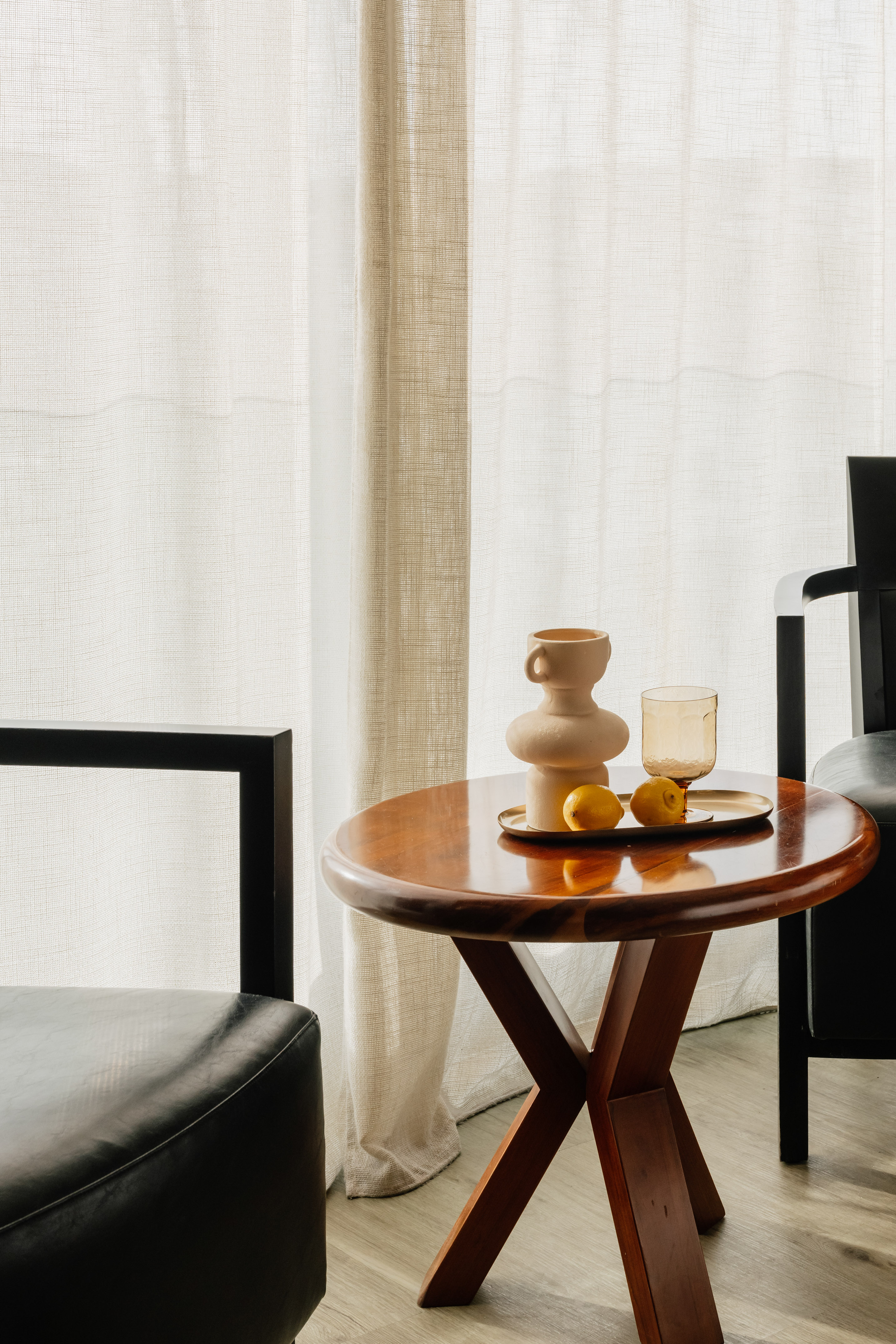 Round wooden coffee table with sculptural vase, lemons, and sheer white curtains.