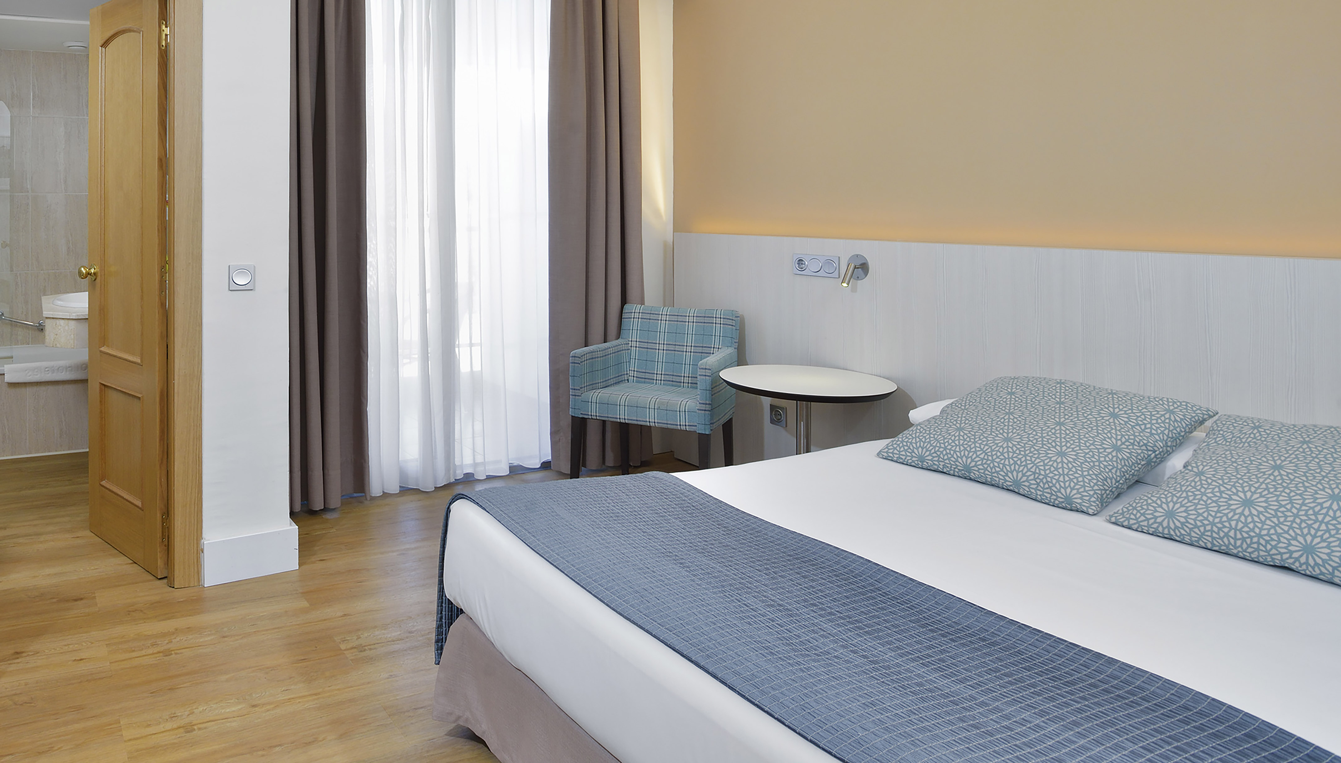 Hotel bedroom with a bed, blue pillows, wooden floor, and window.