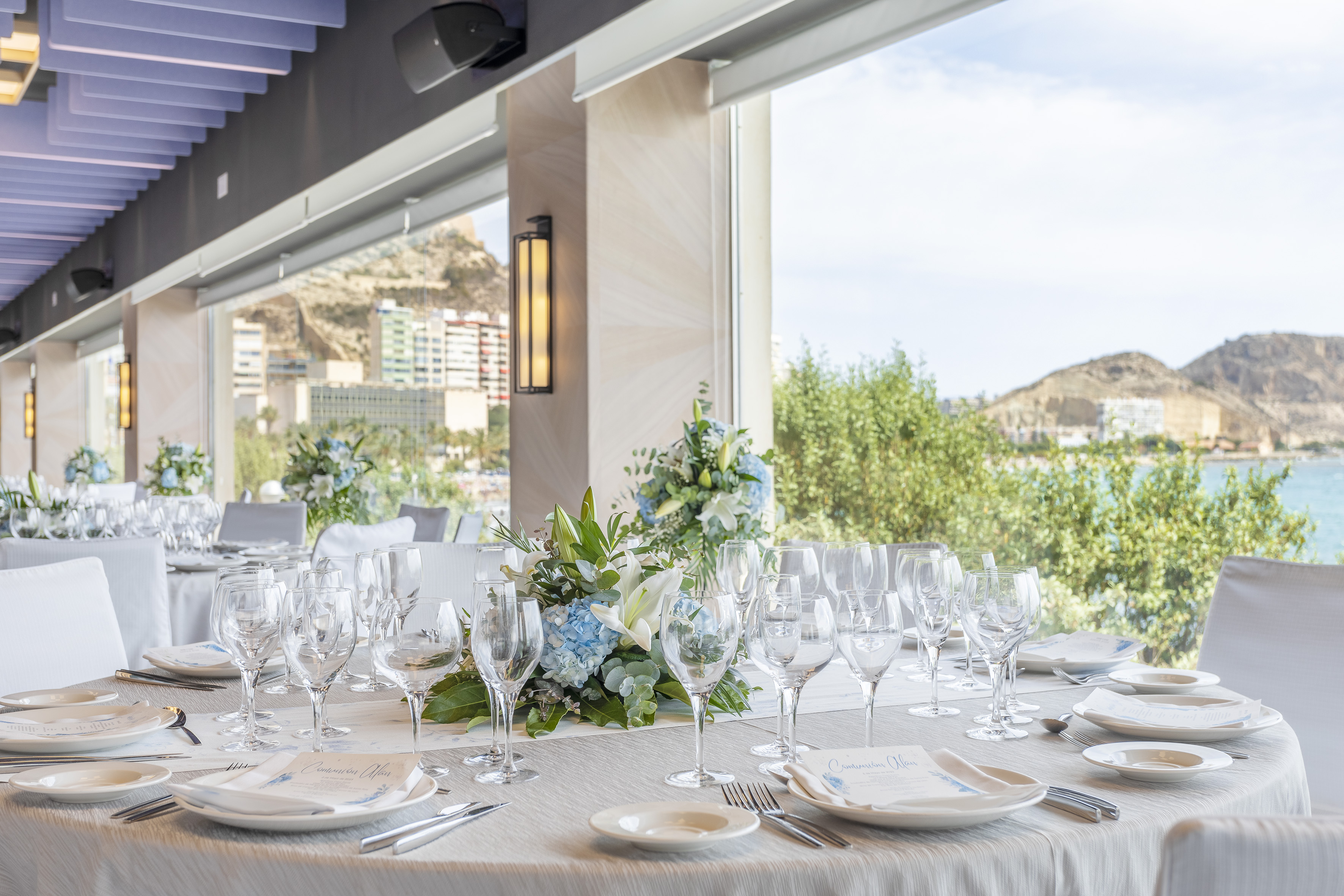 Formal dining tables with flowers, overlooking a coastal bay and mountains.
