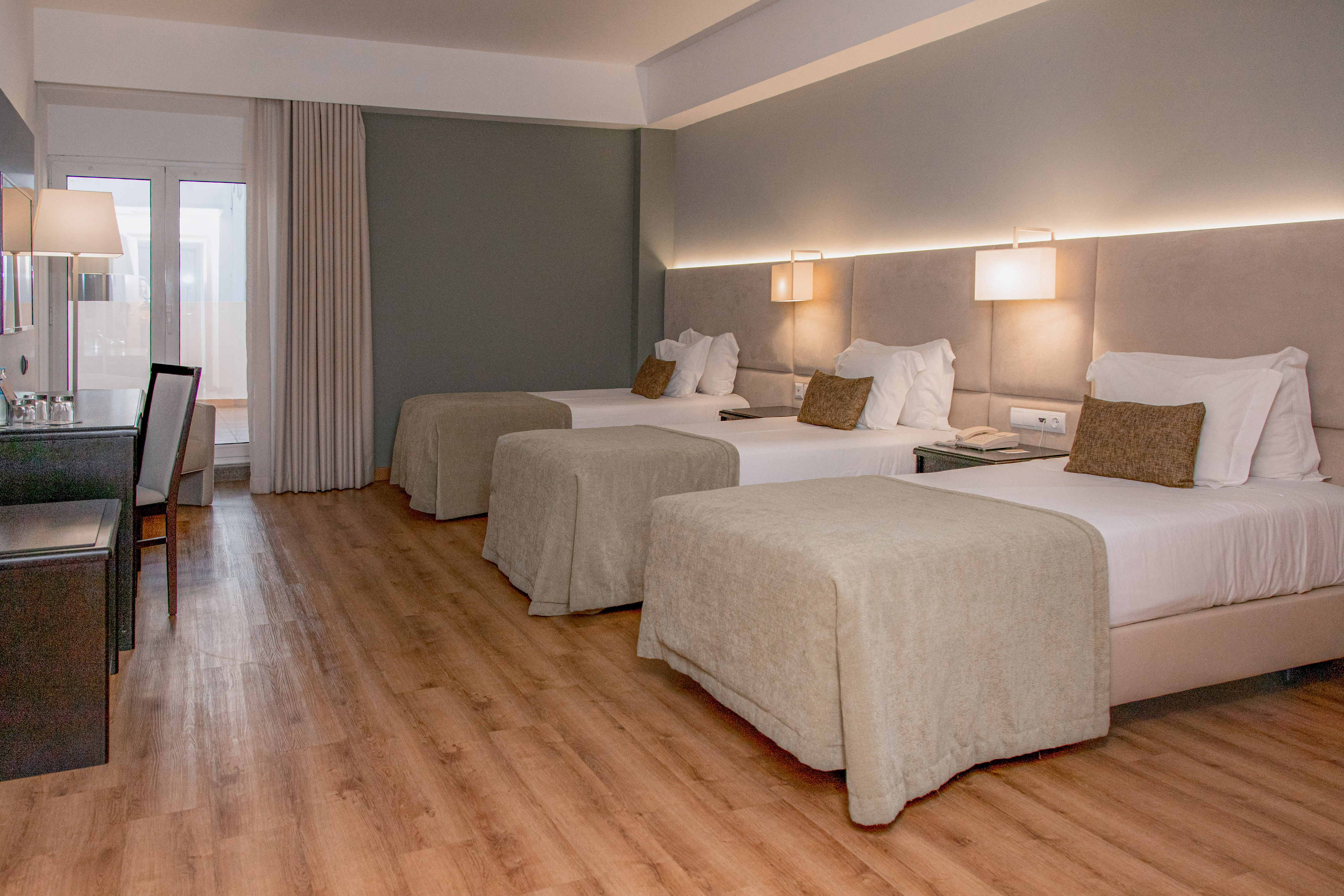 Modern triple hotel room with three single beds, wooden floor, and desk.