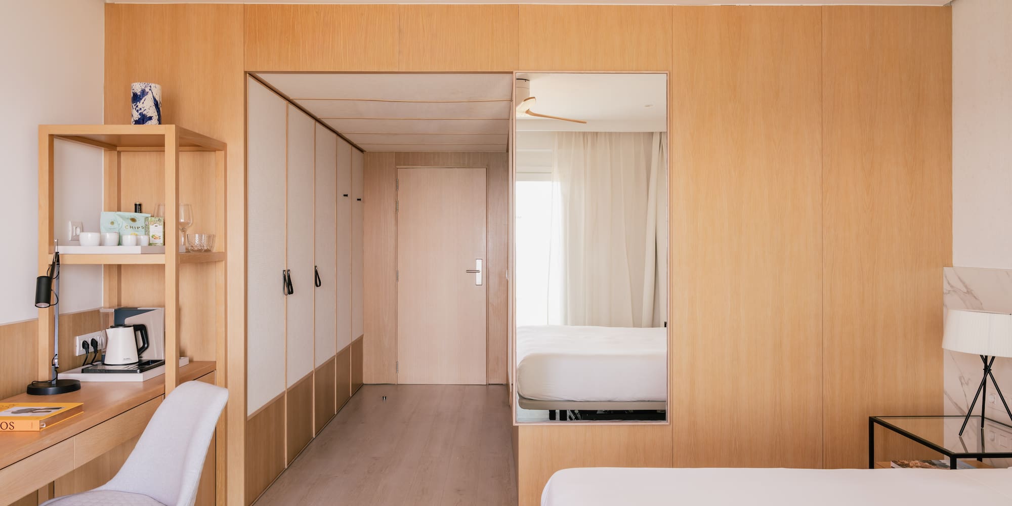 Modern hotel room with wooden walls, desk, white bed, and large mirror.