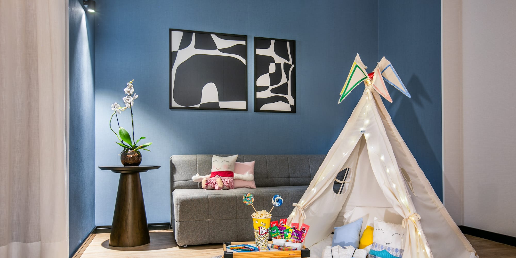 Kids' teepee tent play area with snacks, gray sofa, blue wall, and modern art.