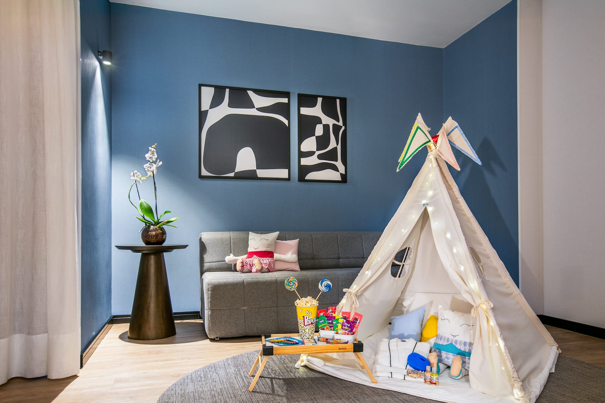 Kids' teepee tent play area with snacks, gray sofa, blue wall, and modern art.