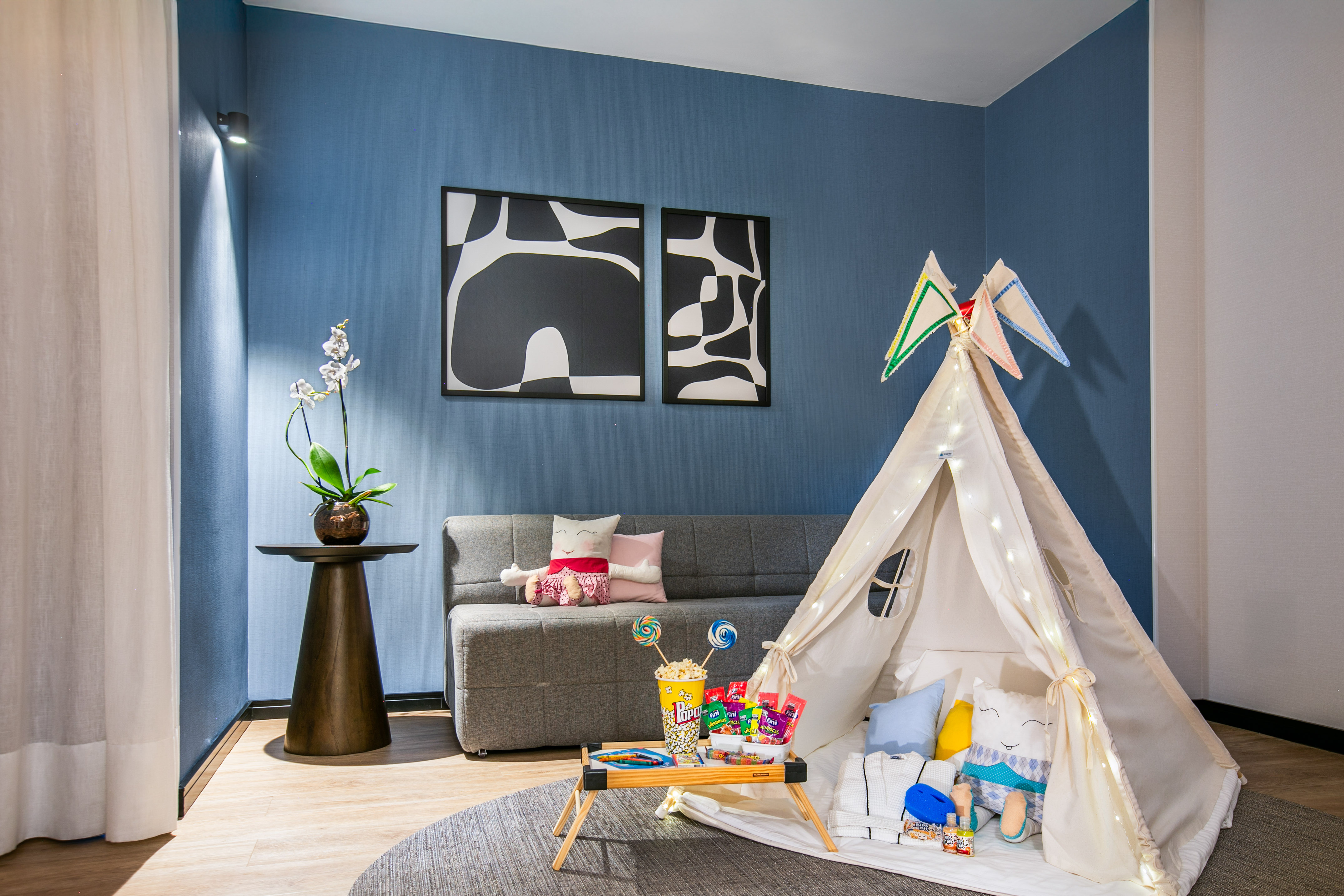 Kids' teepee tent play area with snacks, gray sofa, blue wall, and modern art.