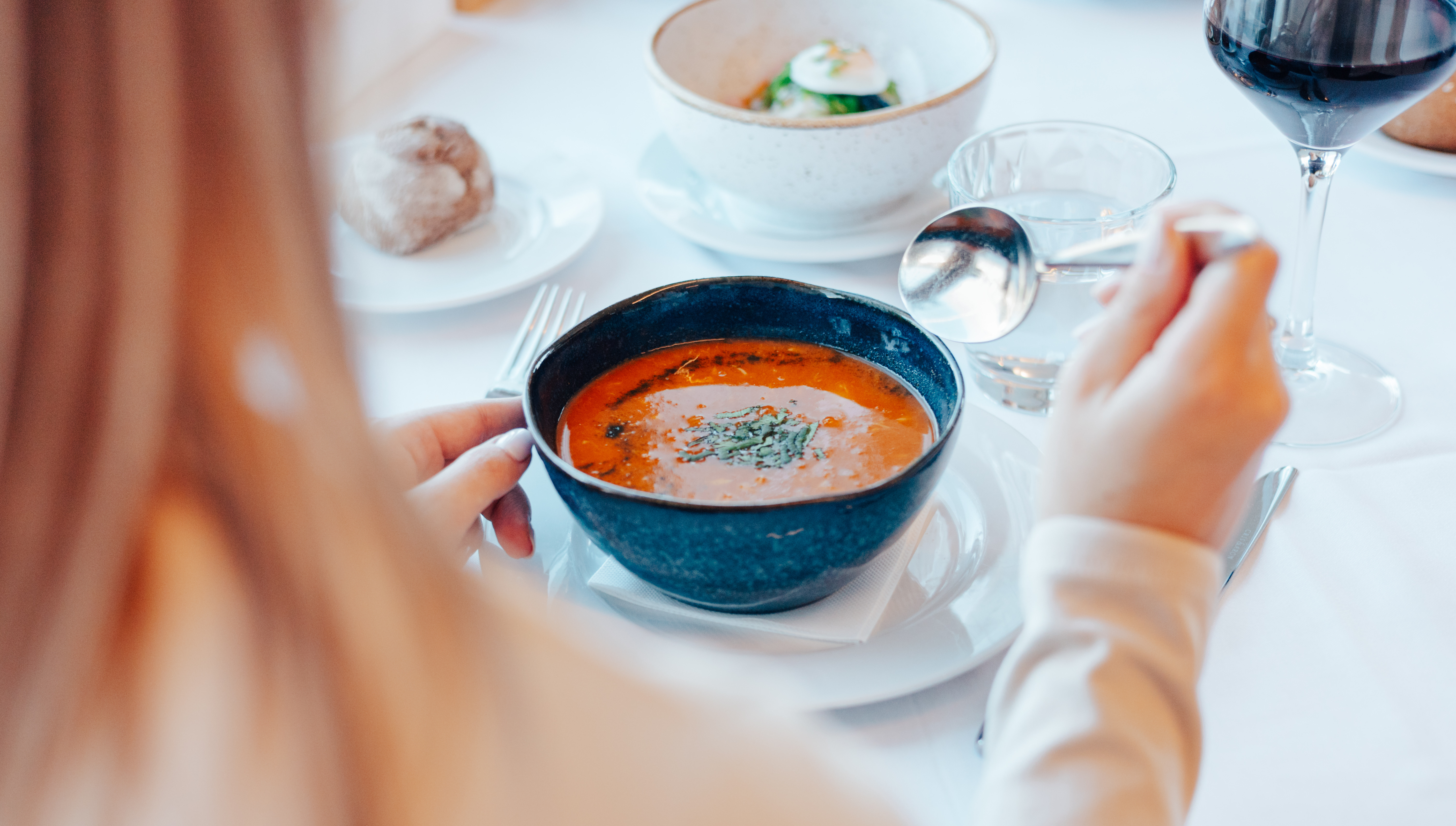 Person eating red soup with herbs, wine, and bread at a restaurant table.