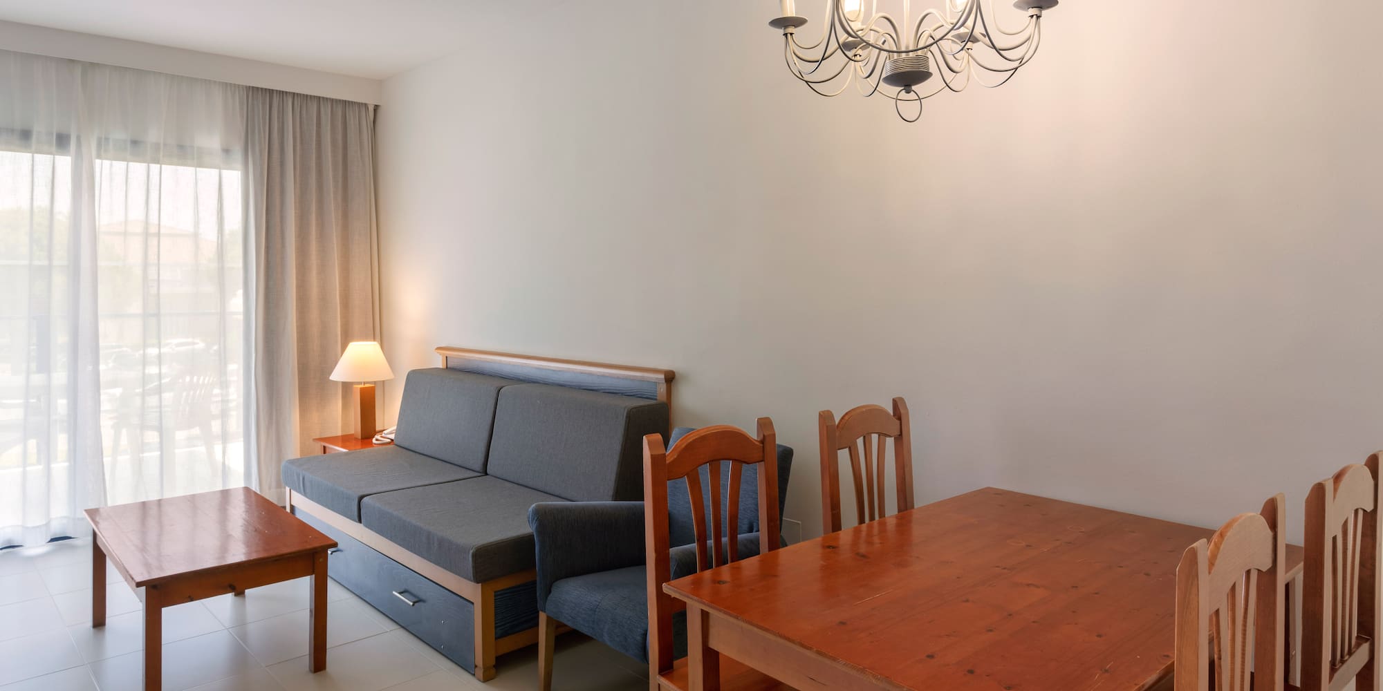 Bright living room with gray sofa bed, wooden dining table, coffee table, and chandelier.
