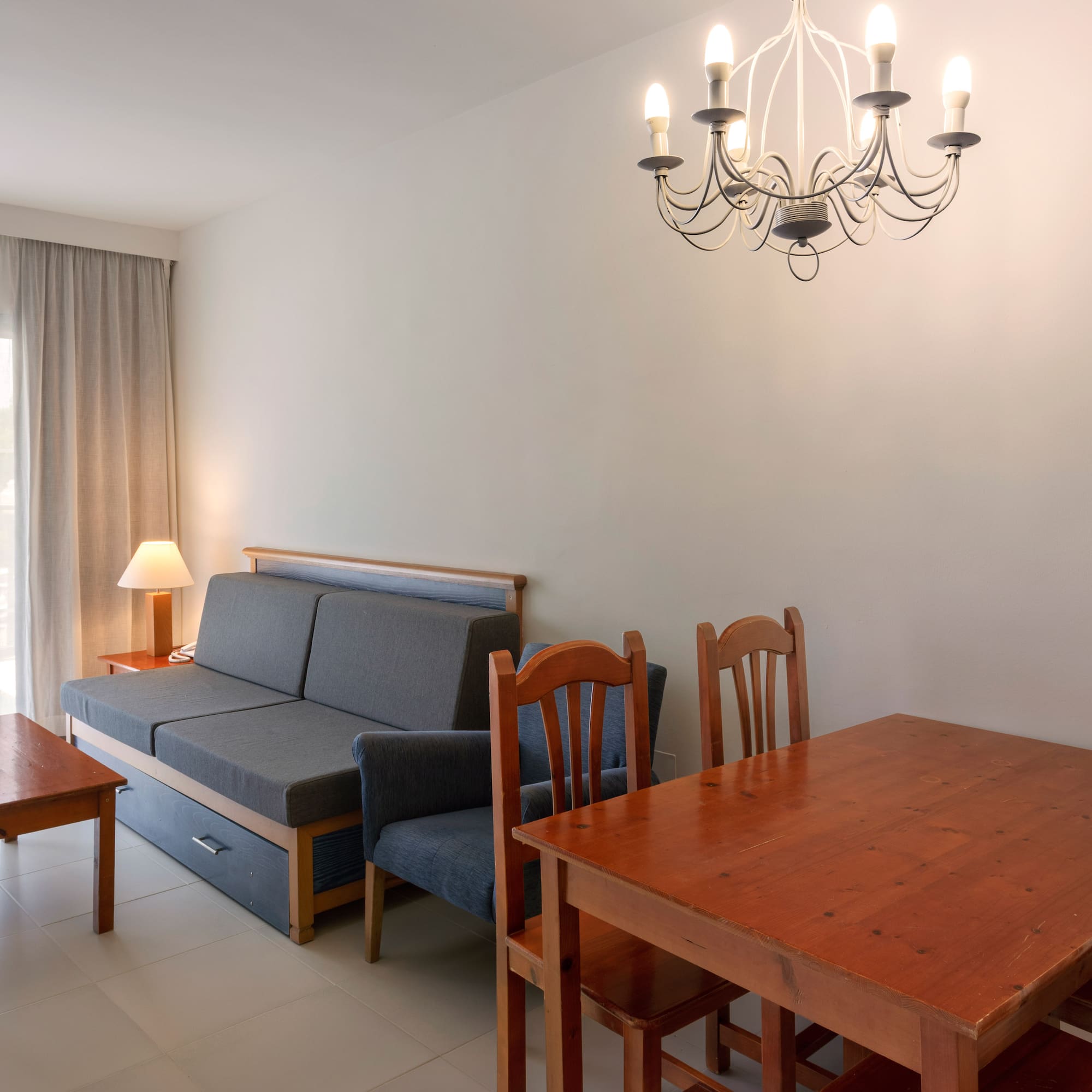 Bright living room with gray sofa bed, wooden dining table, coffee table, and chandelier.