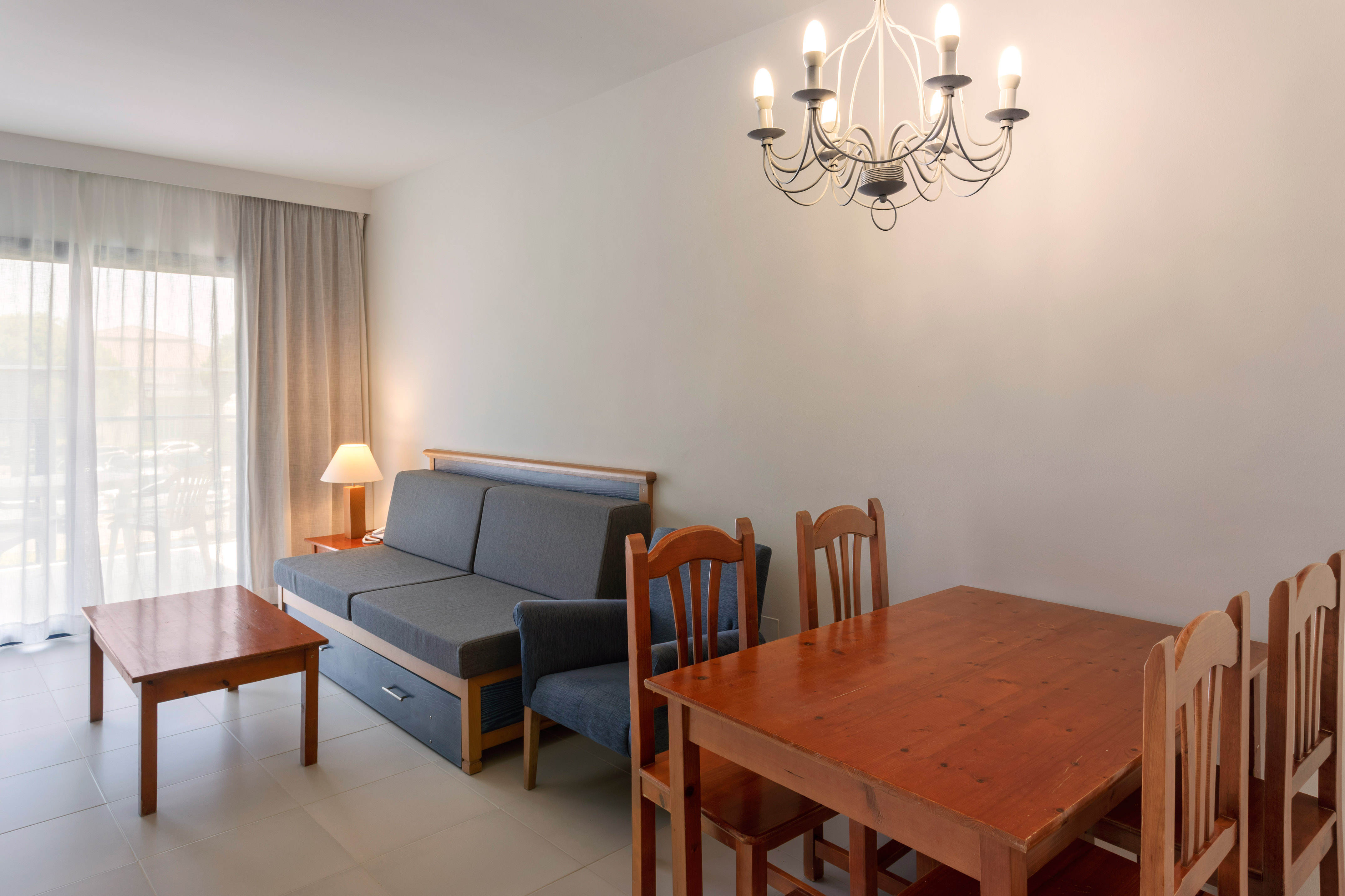Bright living room with gray sofa bed, wooden dining table, coffee table, and chandelier.