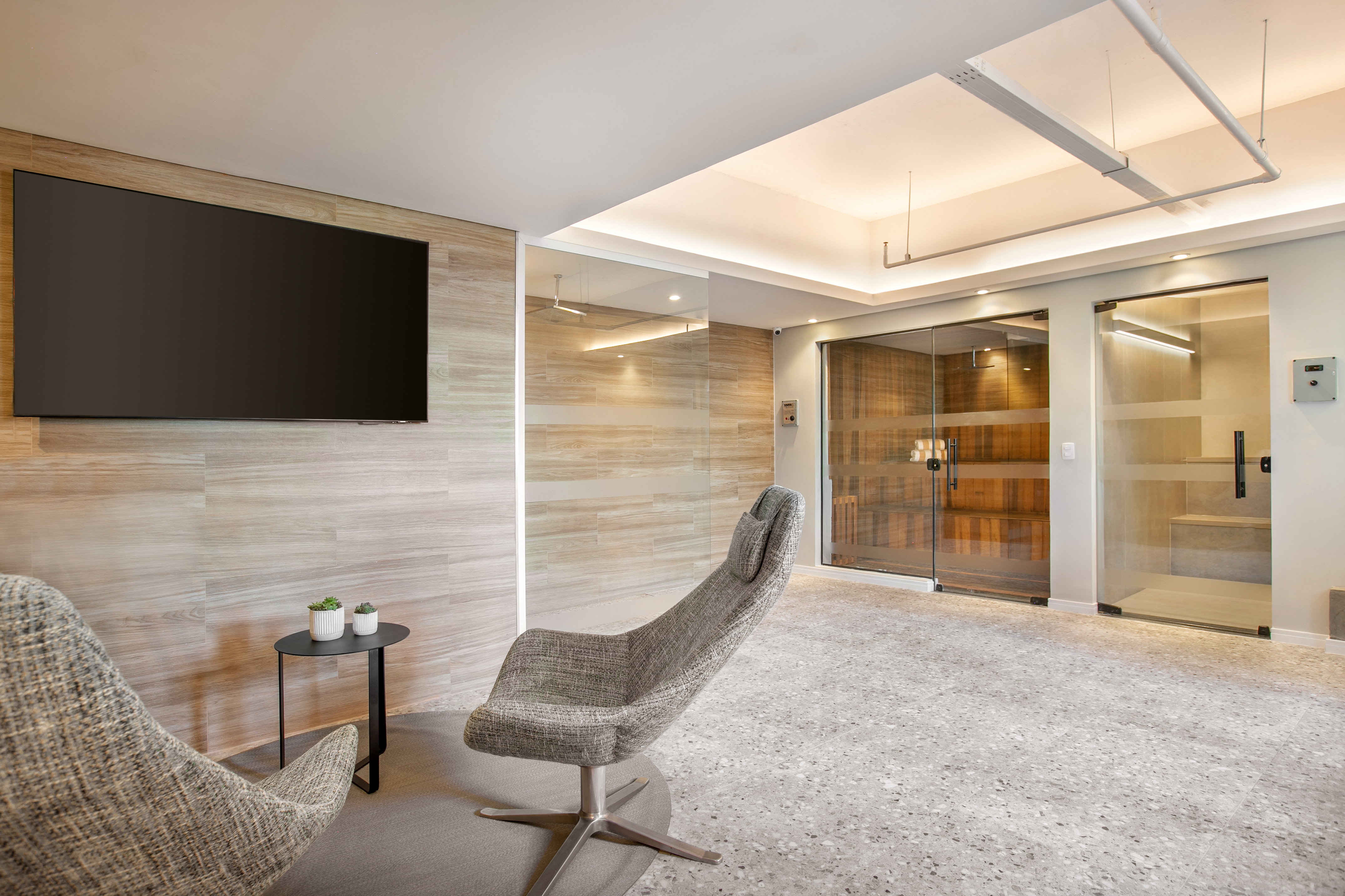 Modern spa lounge with gray chairs, large TV, wood-paneled wall, and sauna entry.